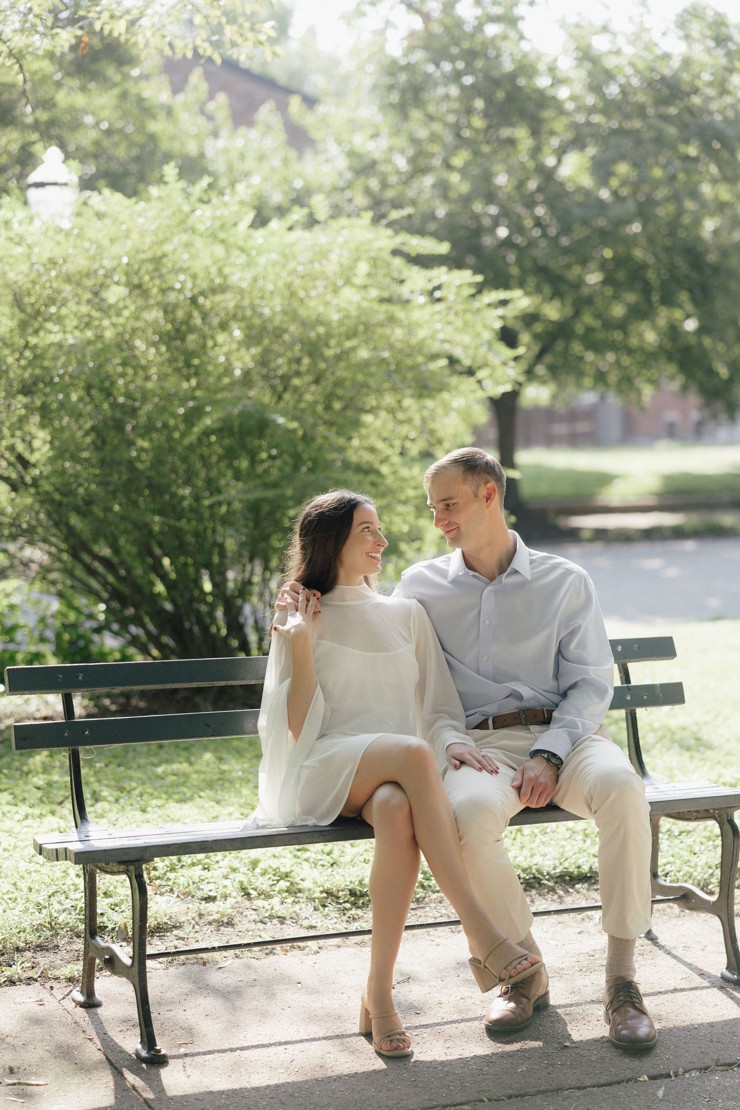 Happy couple sitting close together on a park bench surrounded by lush green trees on a sunny day.