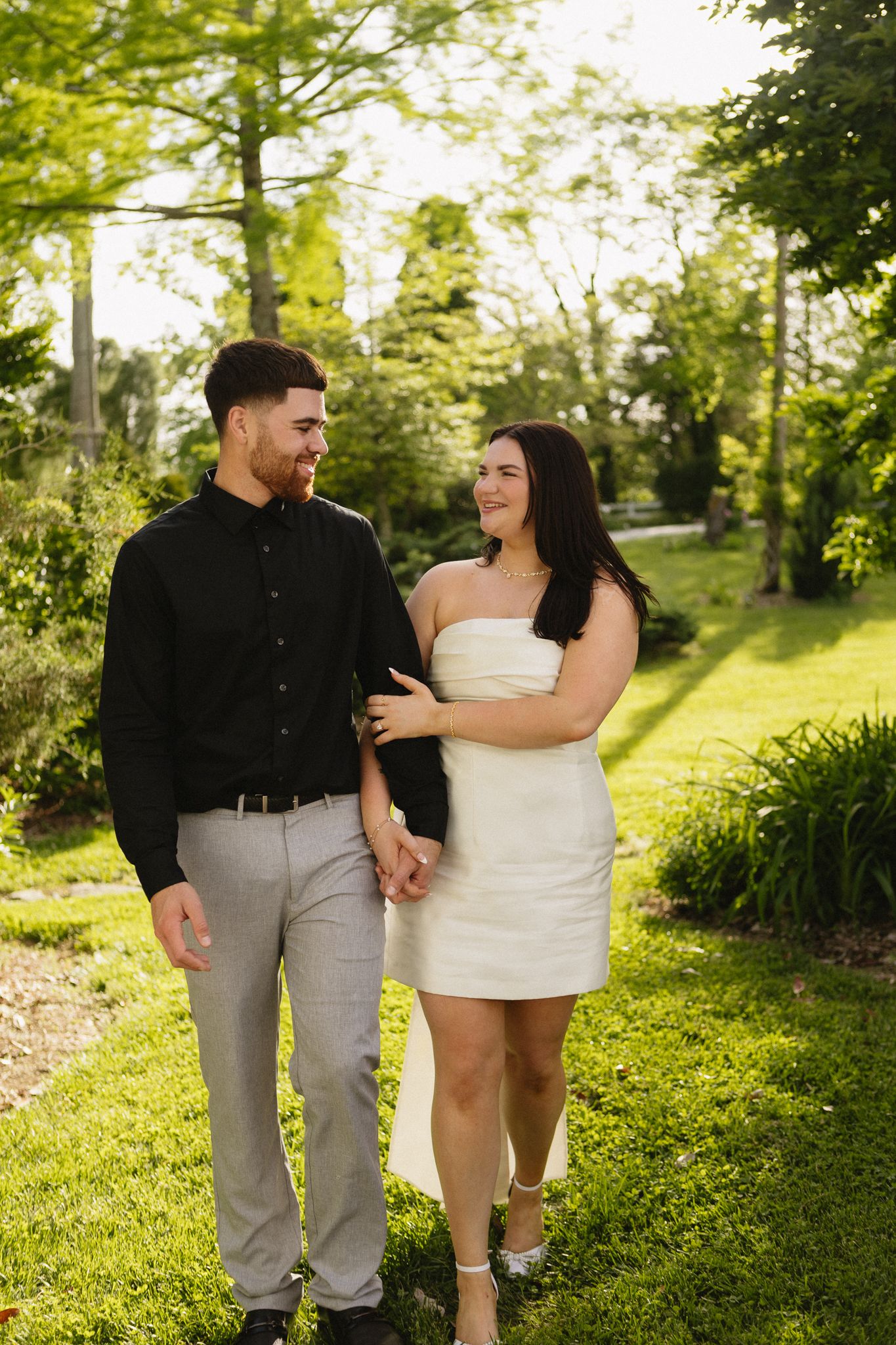 Couple in casual wedding attire walking through a lush green garden path on a sunny summer day.