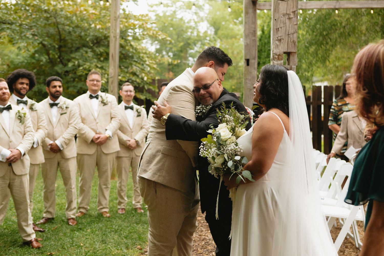 An emotional moment during an outdoor garden wedding ceremony with groomsmen in tan suits standing in the background.