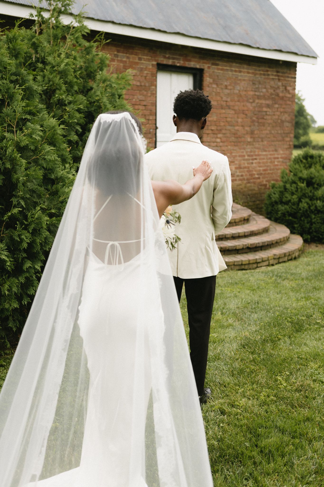 Wedding couple walking on grass path outside brick building with flowing white veil trailing behind.