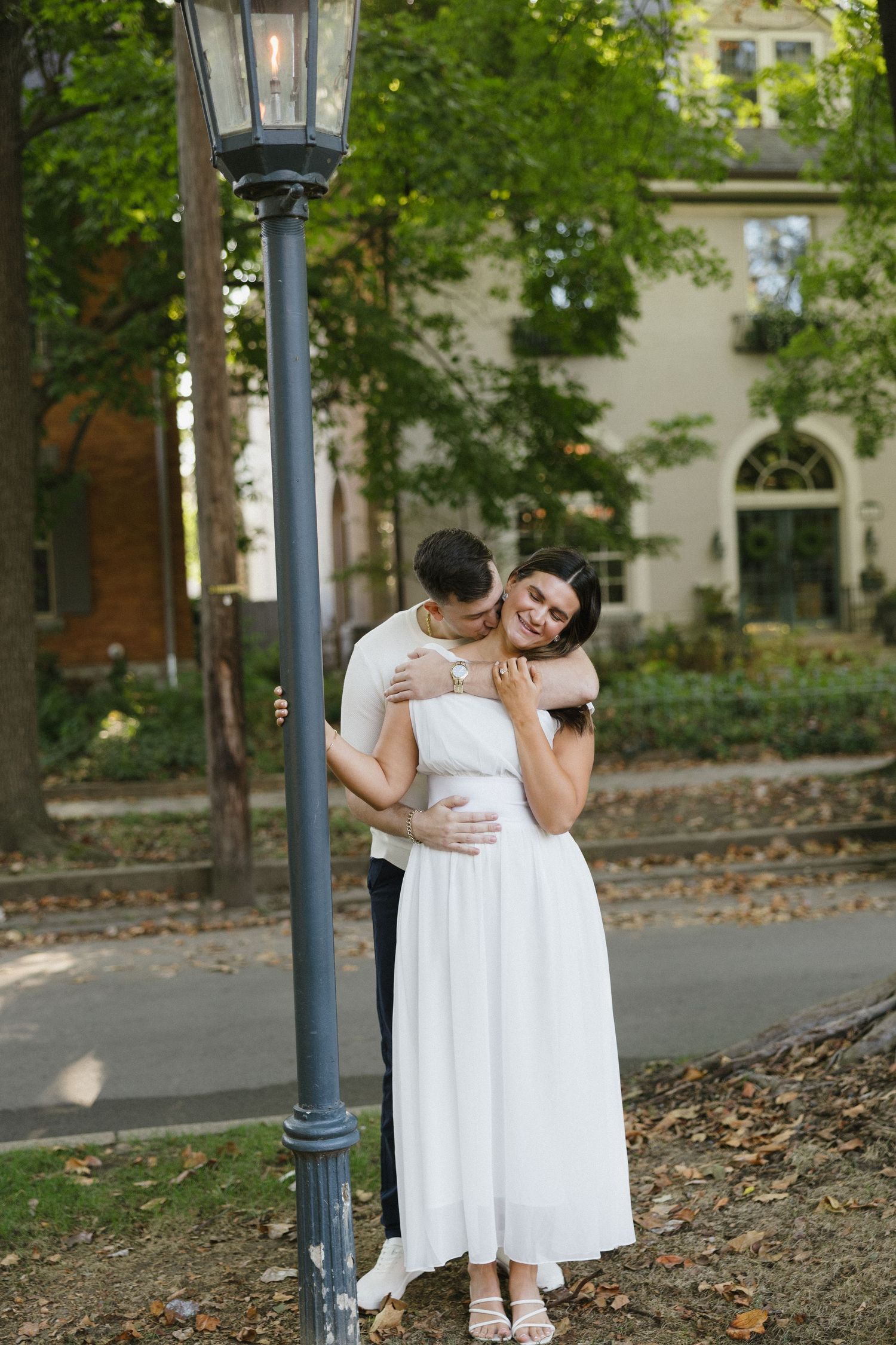 Couple embracing near a vintage street lamp in a lush garden, woman in white dress smiling joyfully.