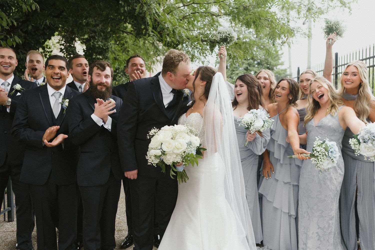 Bride and groom share a kiss surrounded by cheering wedding party in gray bridesmaid dresses and black tuxedos outdoors.