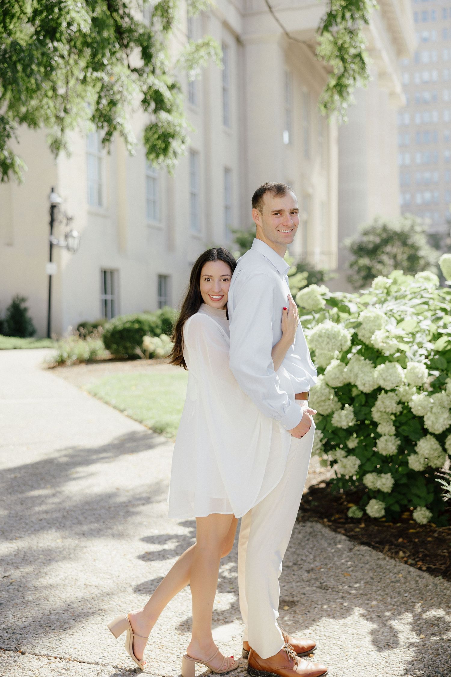 Smiling couple posing together near white hydrangeas outside a classic white building on a sunny day.