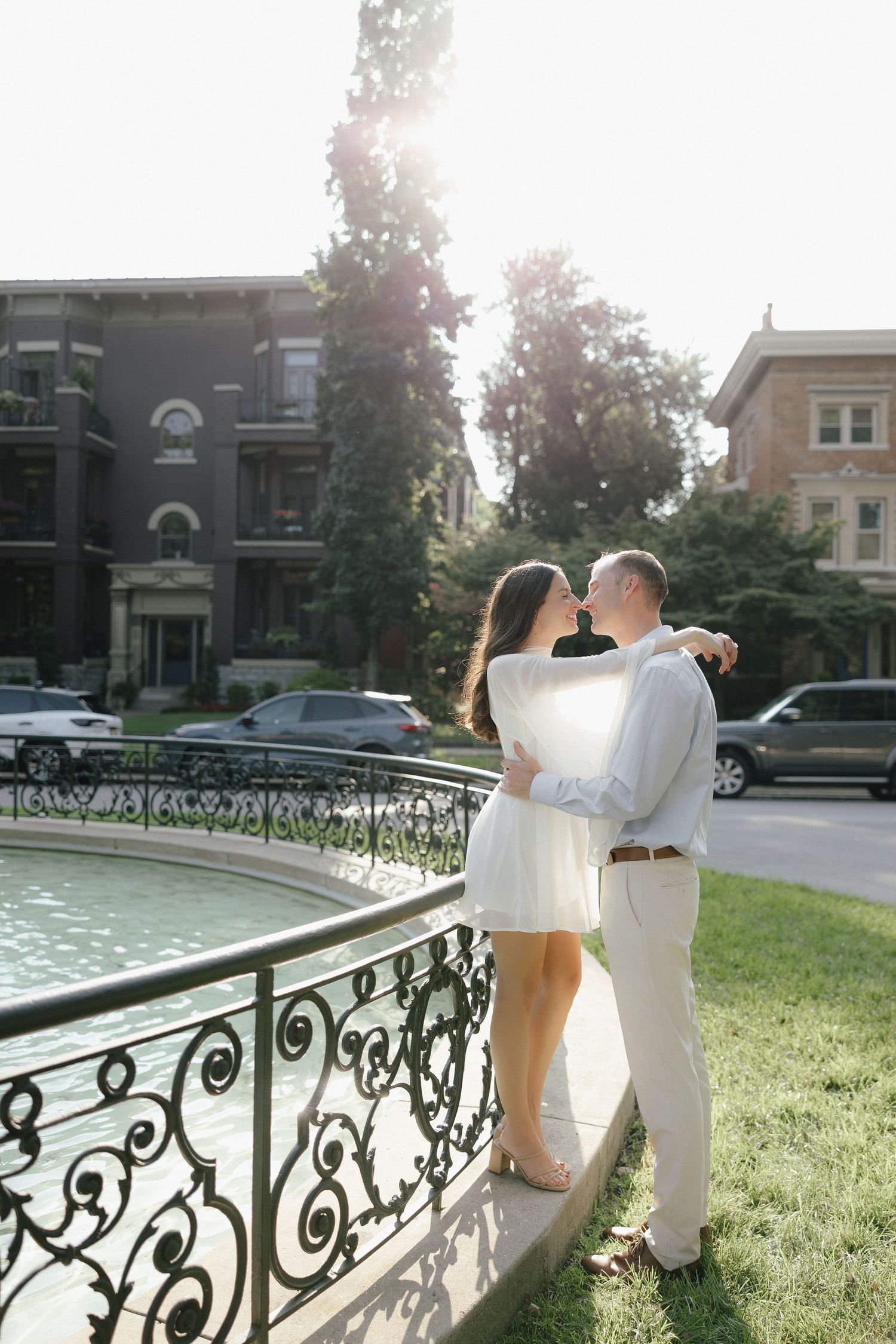 Couple embracing near an ornate fountain with sunlight glowing behind them in an urban park setting.