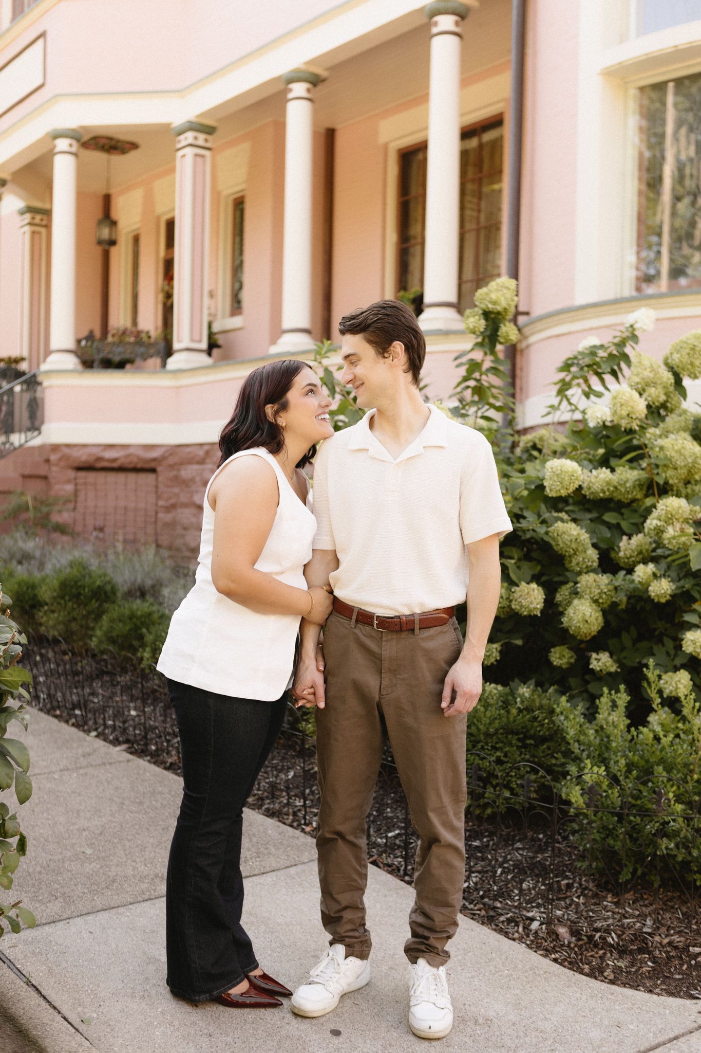 A couple poses together near white columns and hydrangea bushes outside a building.