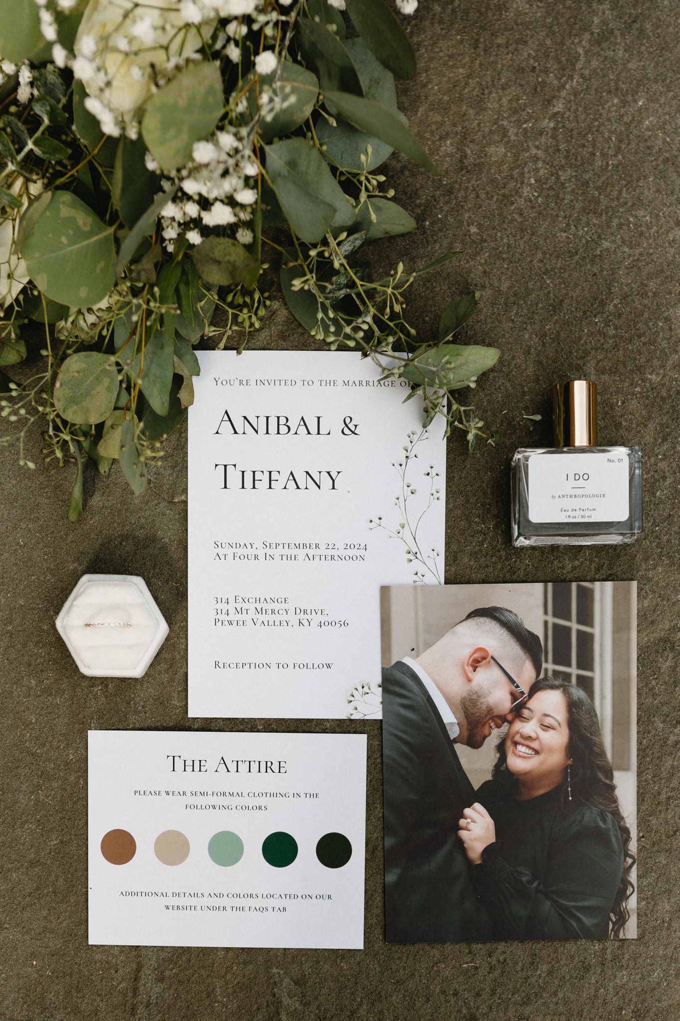 Wedding invitation flatlay with foliage, invitation card, color palette, and couple photo against grey background.