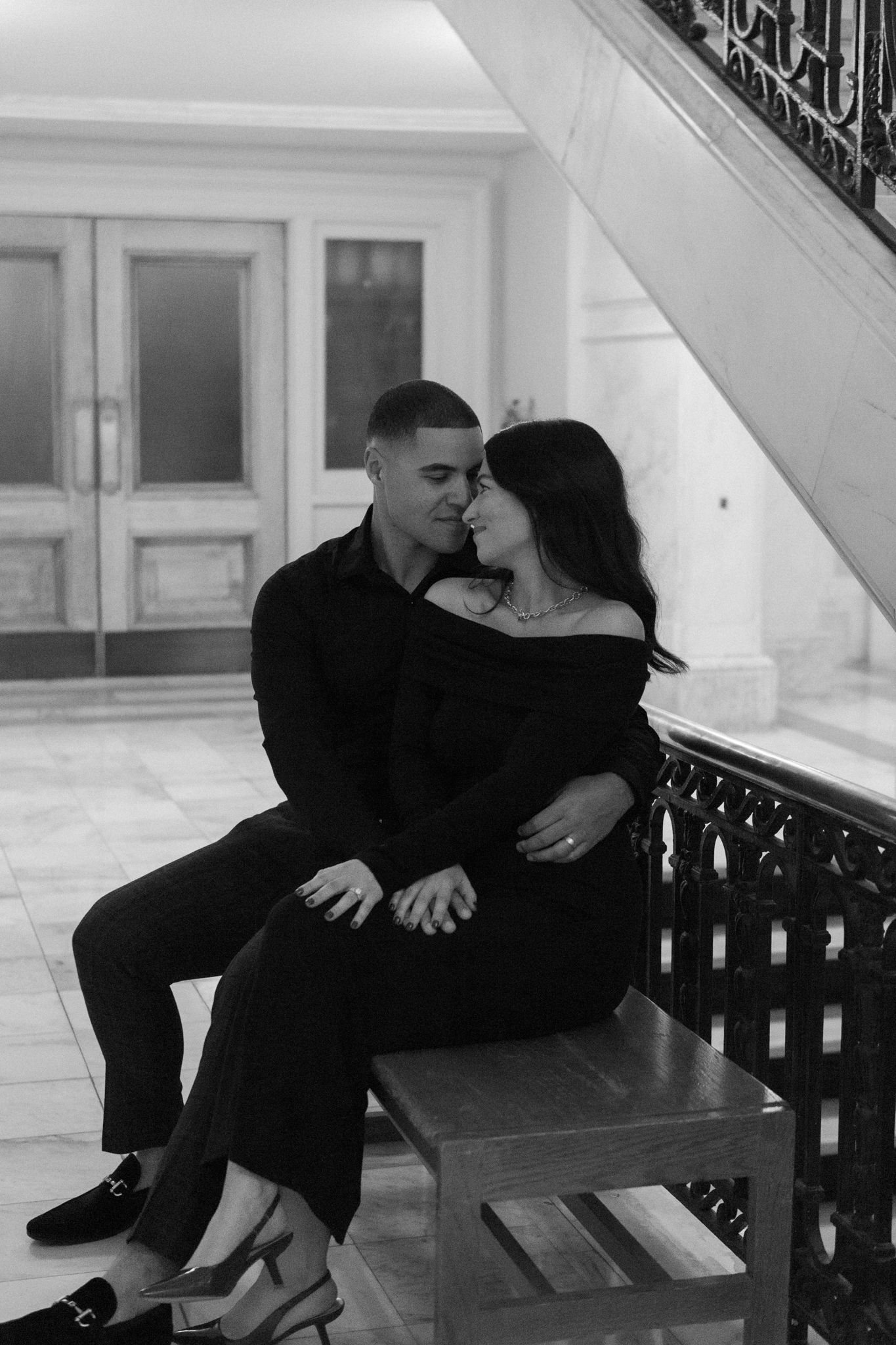 A romantic black and white portrait of a couple in dark formal attire embracing on an elegant staircase.