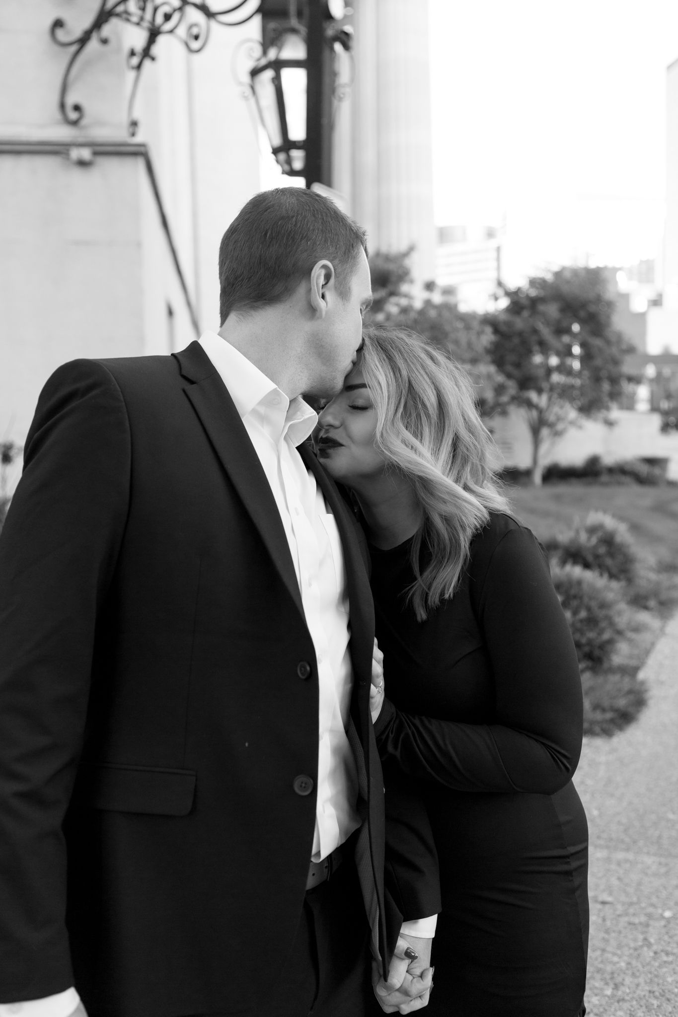 A romantic black and white portrait of a couple in formal attire sharing an intimate moment outdoors.