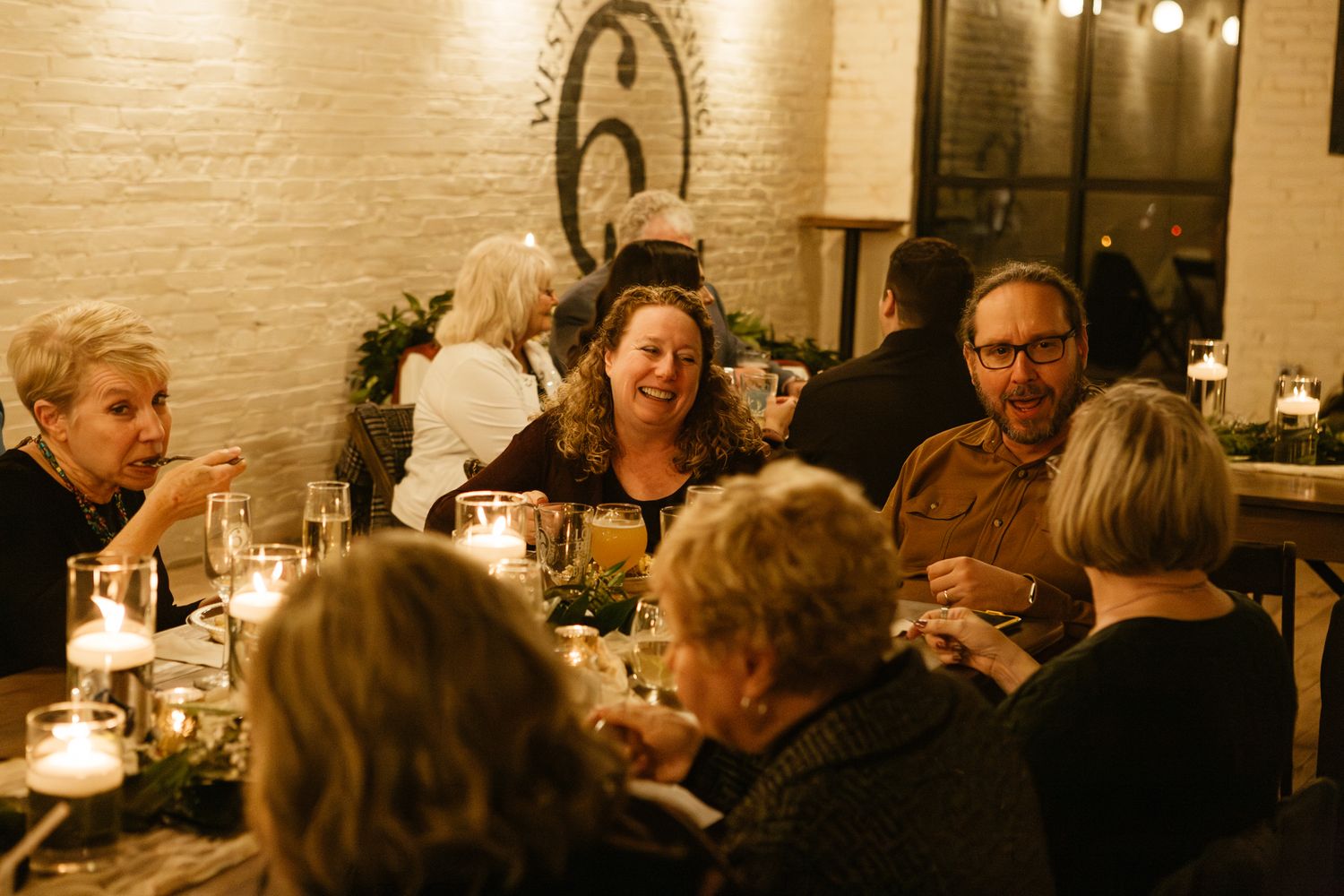 Guests enjoying dinner at a candlelit reception table in a rustic brick-walled venue.