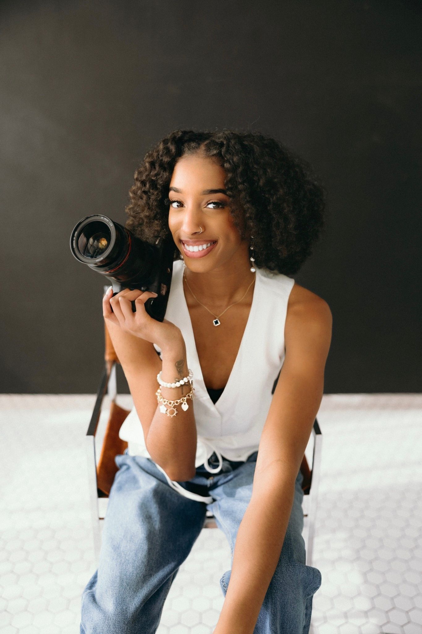 Photographer in white sleeveless top and jeans holds professional camera while sitting and smiling.