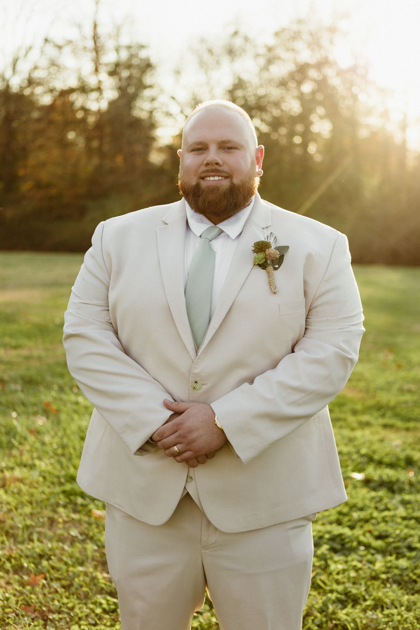 A groom in a cream-colored suit poses in a sunlit field during golden hour with a boutonniere pinned to his lapel.