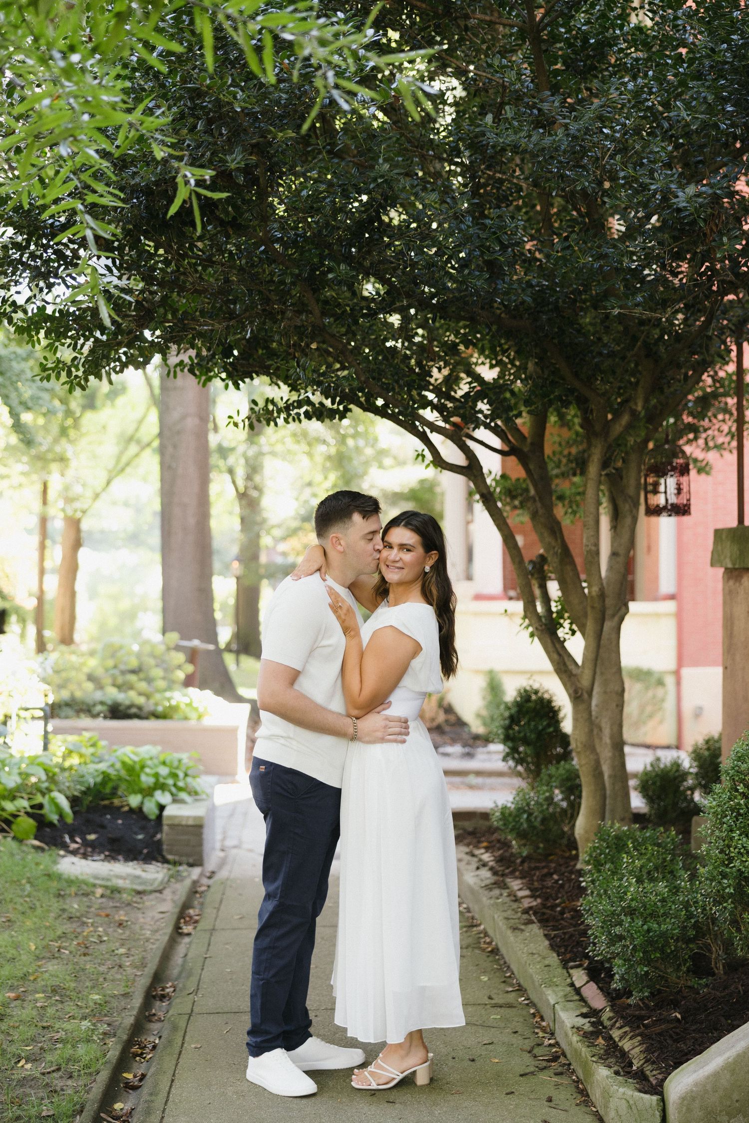 Man kissing woman on the cheek while embracing on a scenic garden walkway lined with trees.