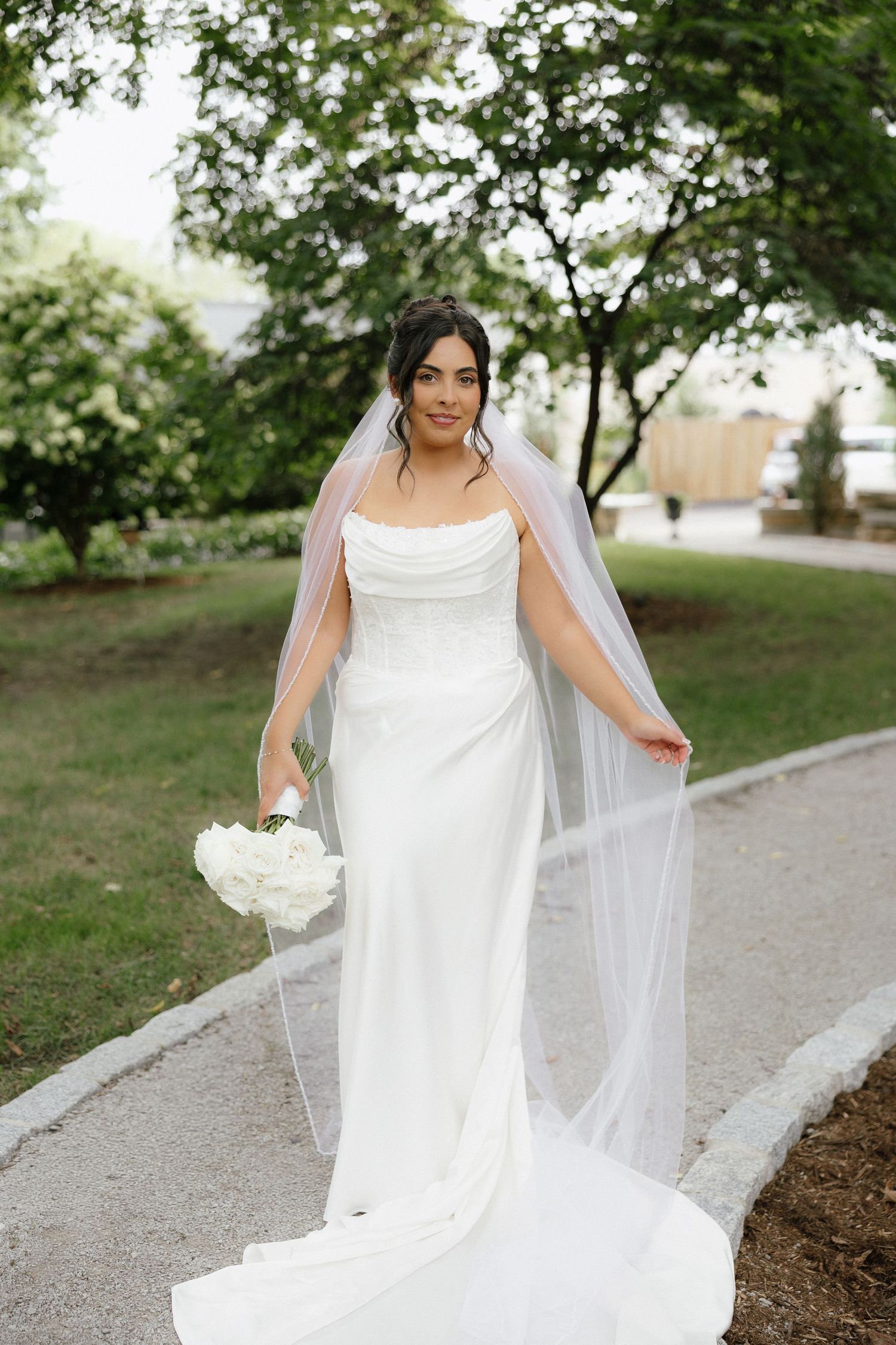 Radiant bride in fitted white gown holding bouquet with long flowing veil in lush garden setting.