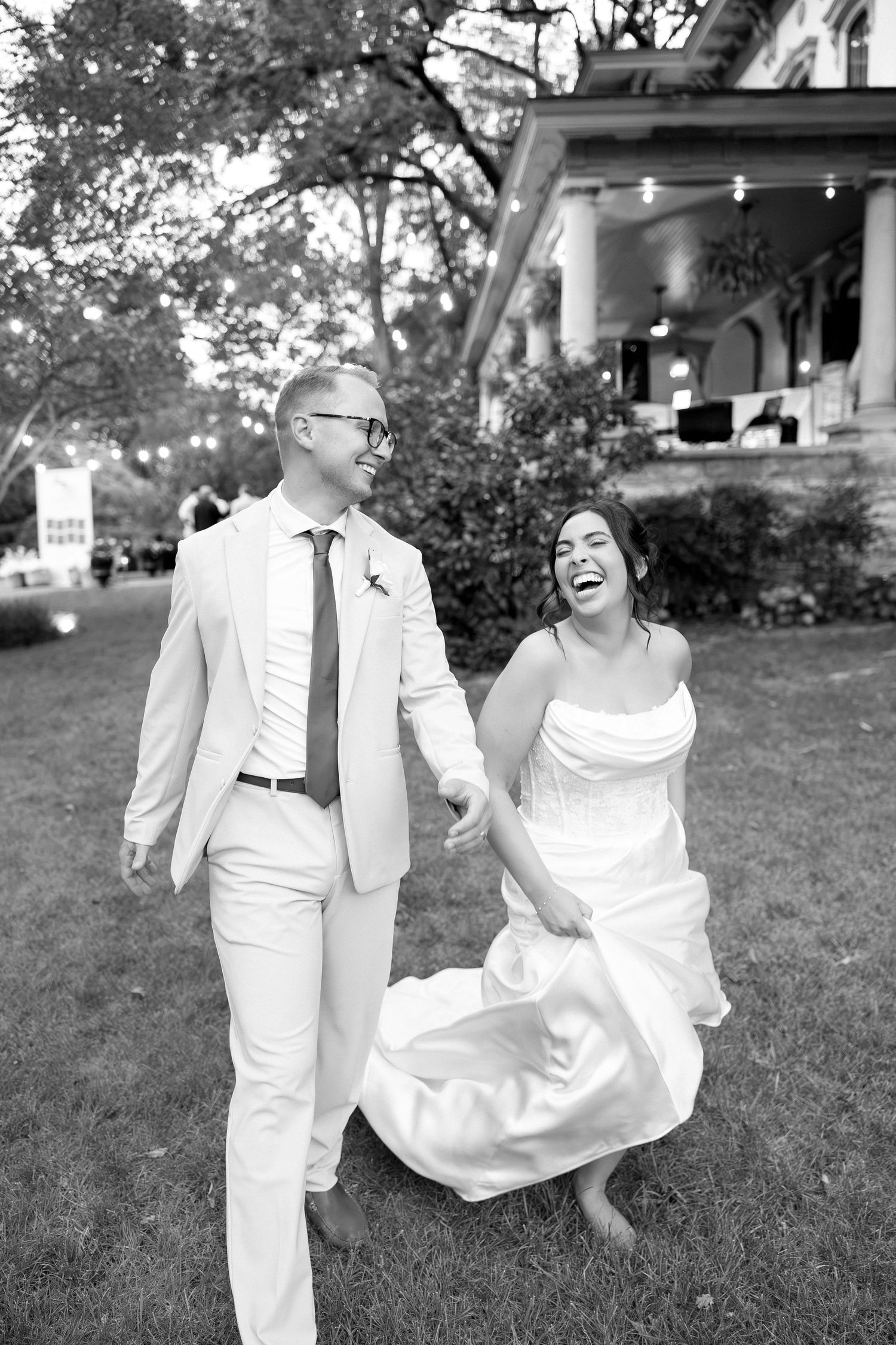Joyful bride and groom in black and white photo laughing while running across lawn at outdoor wedding.