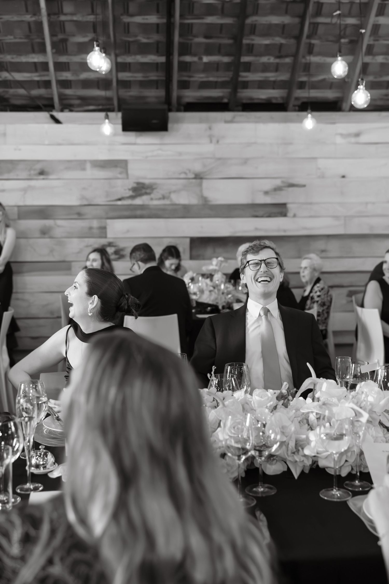 Black and white photo of wedding guest laughing joyfully at reception dinner table with string lights.