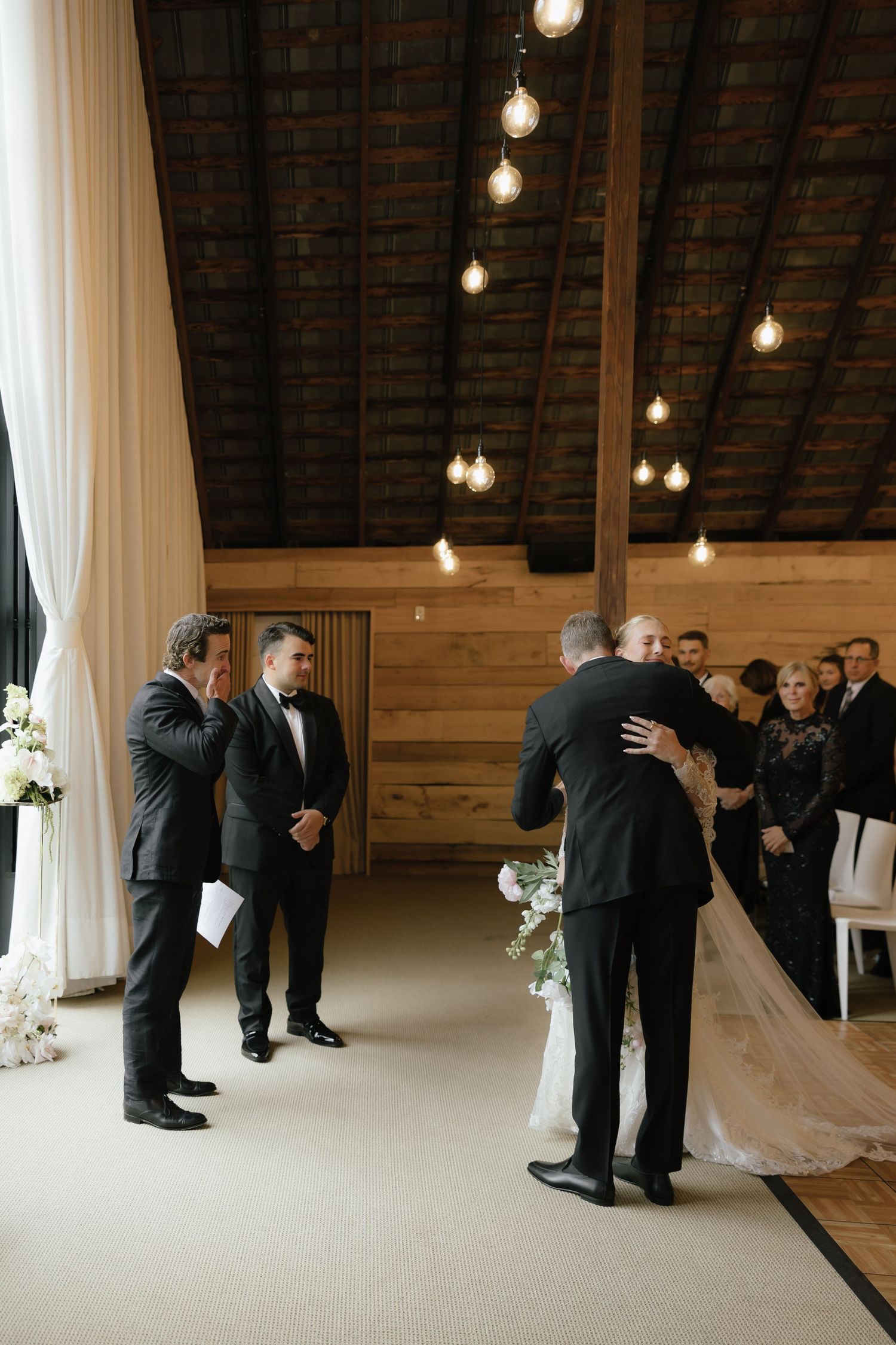 Father walks bride down the aisle at elegant barn wedding ceremony with Edison bulb lighting.