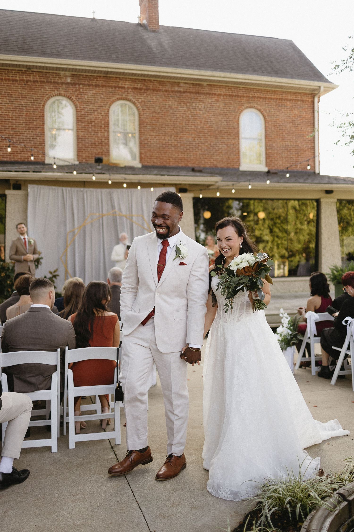 Wedding celebration outside brick venue with white chairs and decorative lighting.