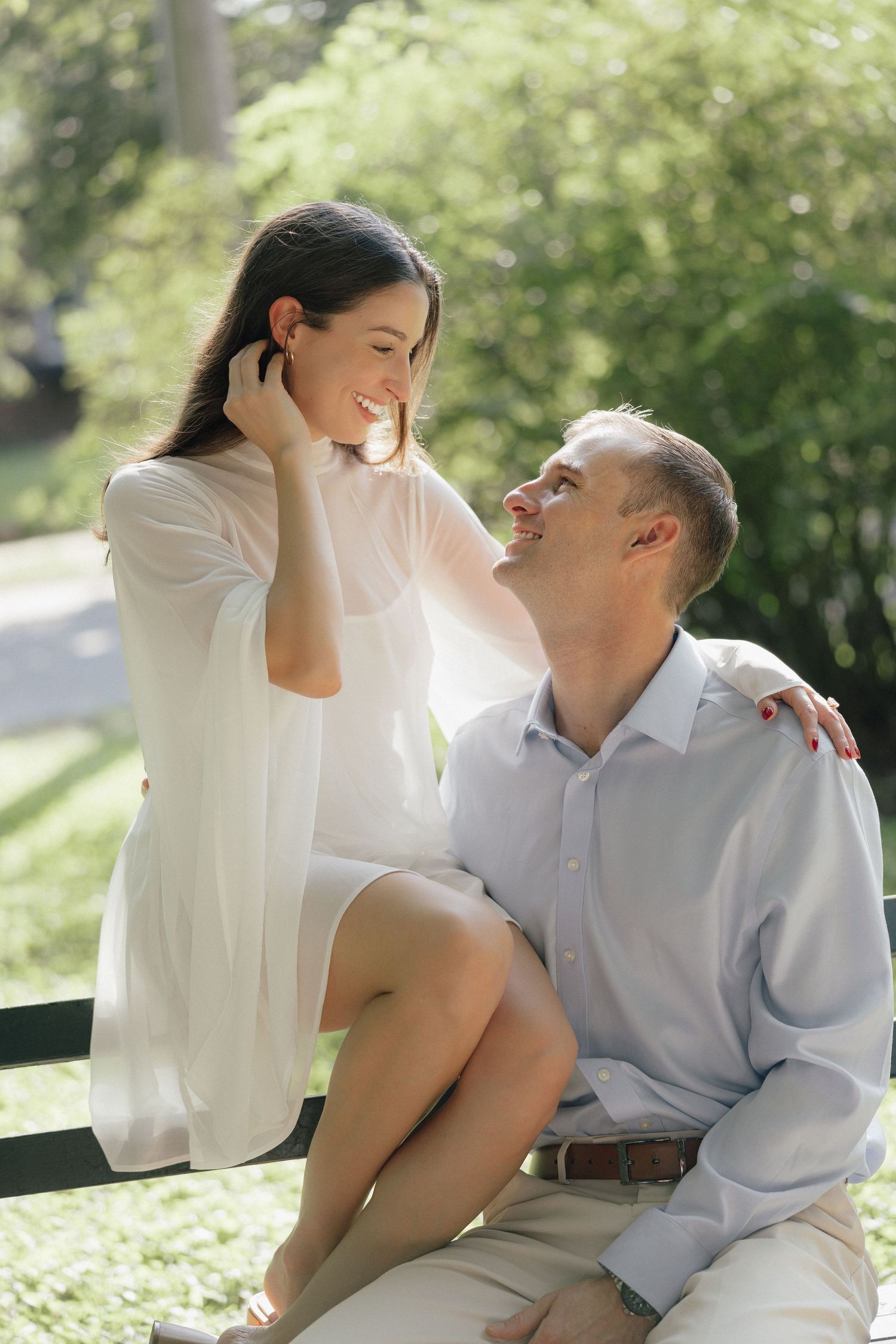 A smiling couple shares a romantic moment on a park bench, woman in white dress sitting while man gazes up at her.