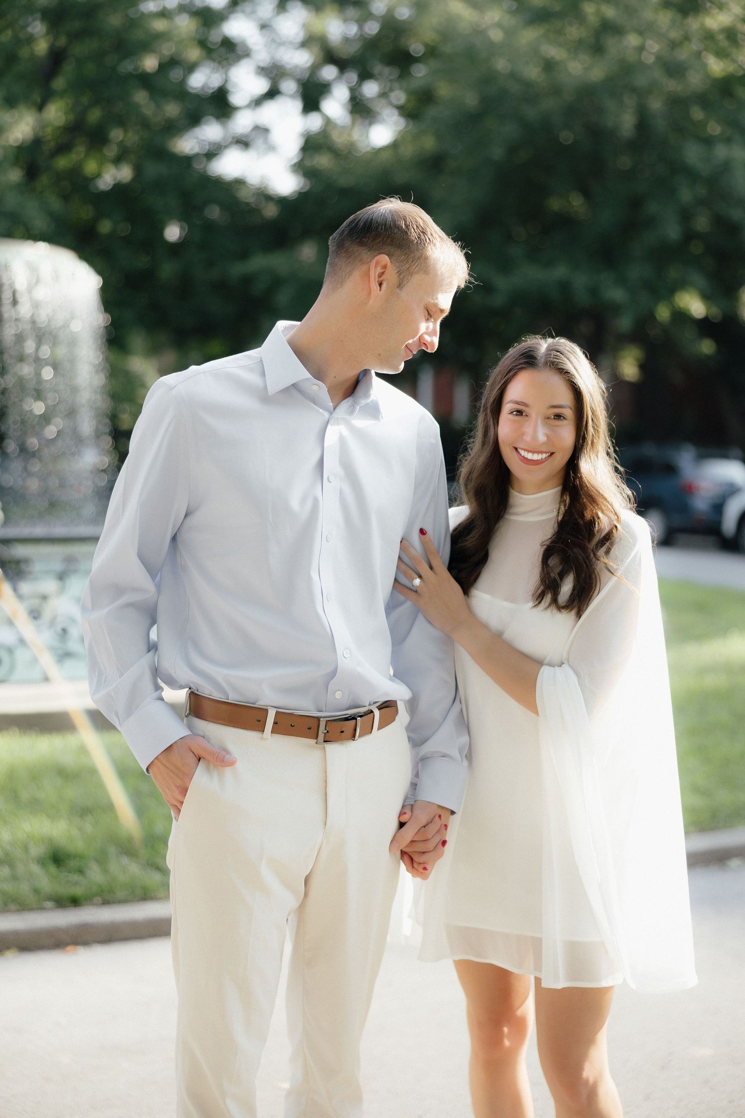 Couple holding hands near a fountain, woman in white dress smiling, man in light blue shirt and cream pants.