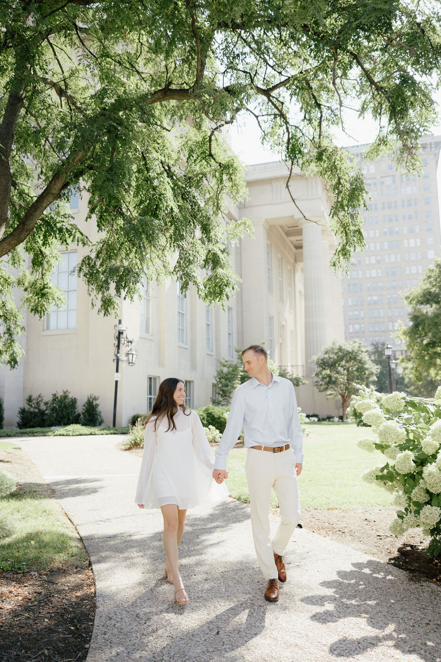 Couple in white outfits holding hands walking along a garden path with blooming hydrangeas and a grand building backdrop.