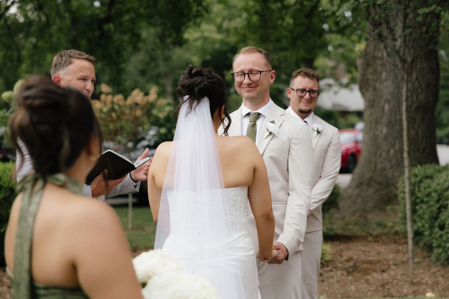 Groom in beige suit smiles at bride during outdoor wedding ceremony with officiant and groomsman.