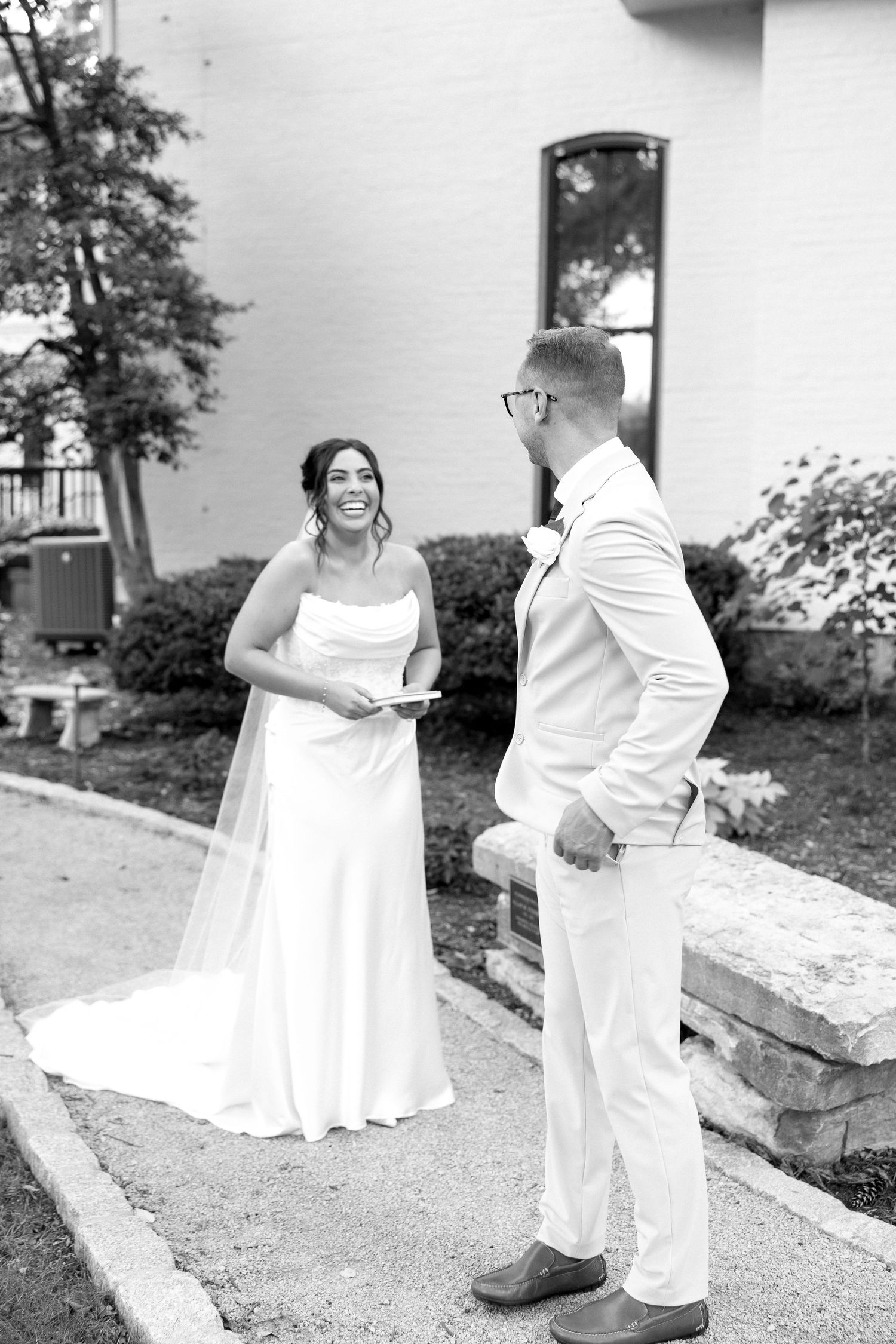 Bride and groom share a joyful first look moment outside a white building.