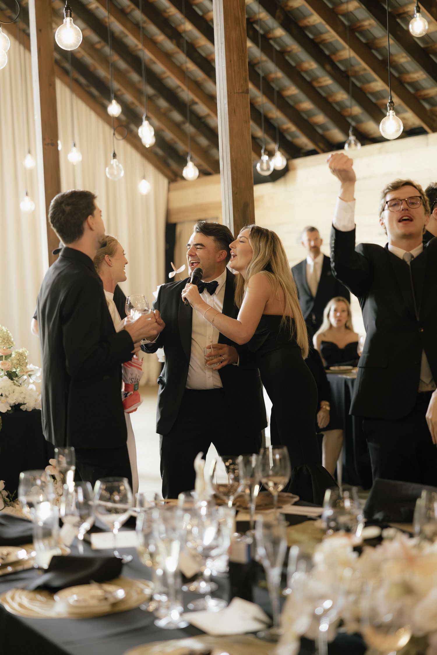 Wedding guests in black tie celebrating with groom in tuxedo under Edison bulb lights in barn reception.