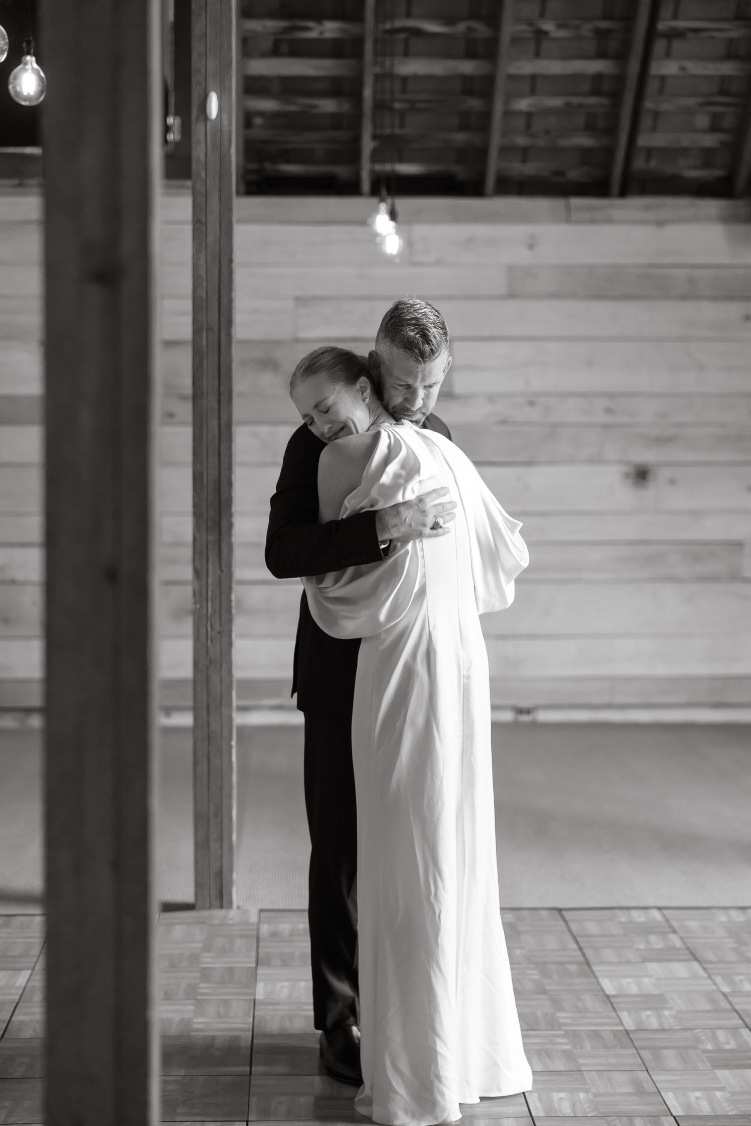 Bride and groom share tender embrace during first dance in rustic wooden barn wedding venue.