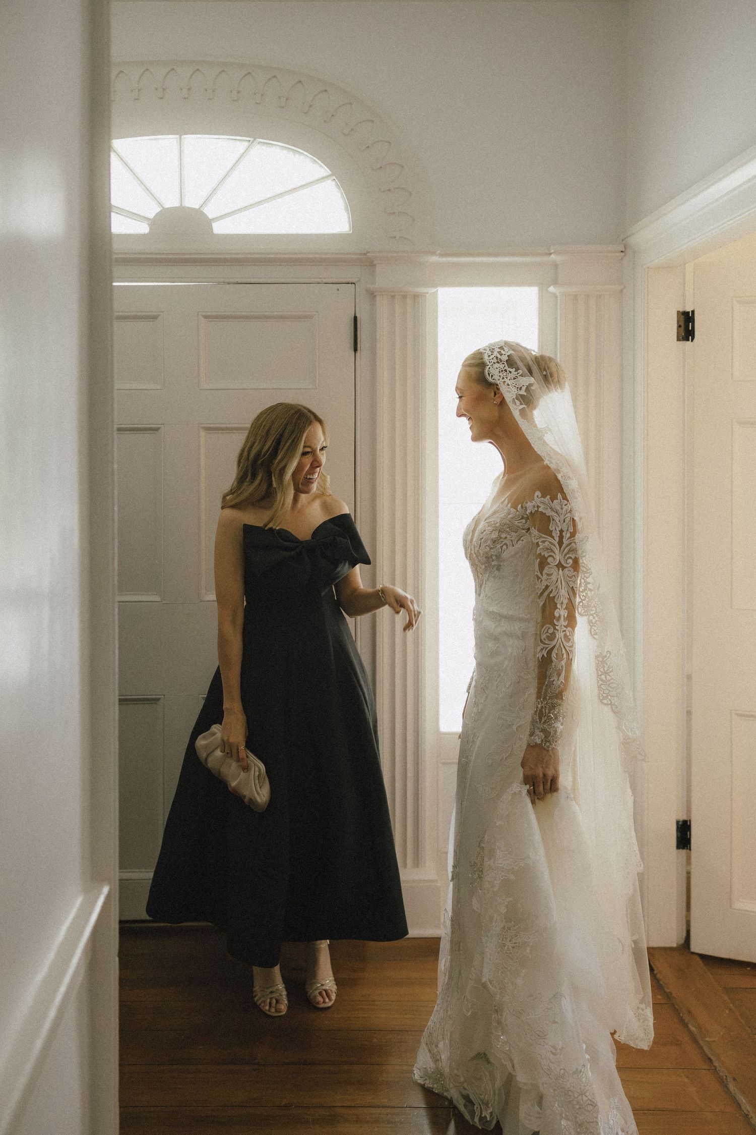 Bride in lace wedding gown with veil stands with bridesmaid in black off-shoulder dress in elegant white hallway.