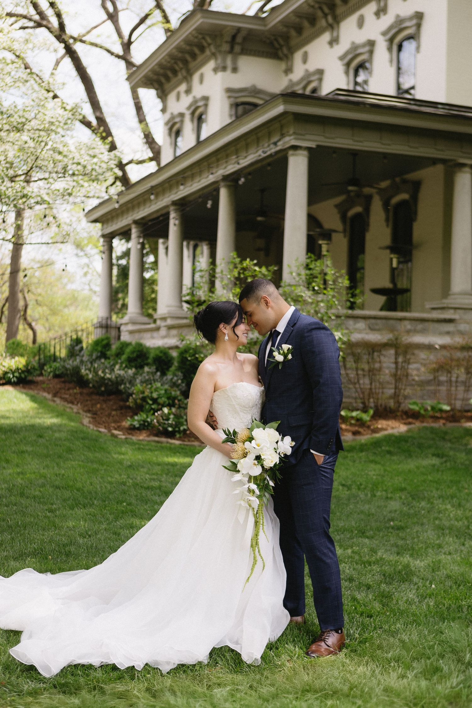 Bride and groom share a romantic moment on lush green lawn before elegant columned mansion on their wedding day.