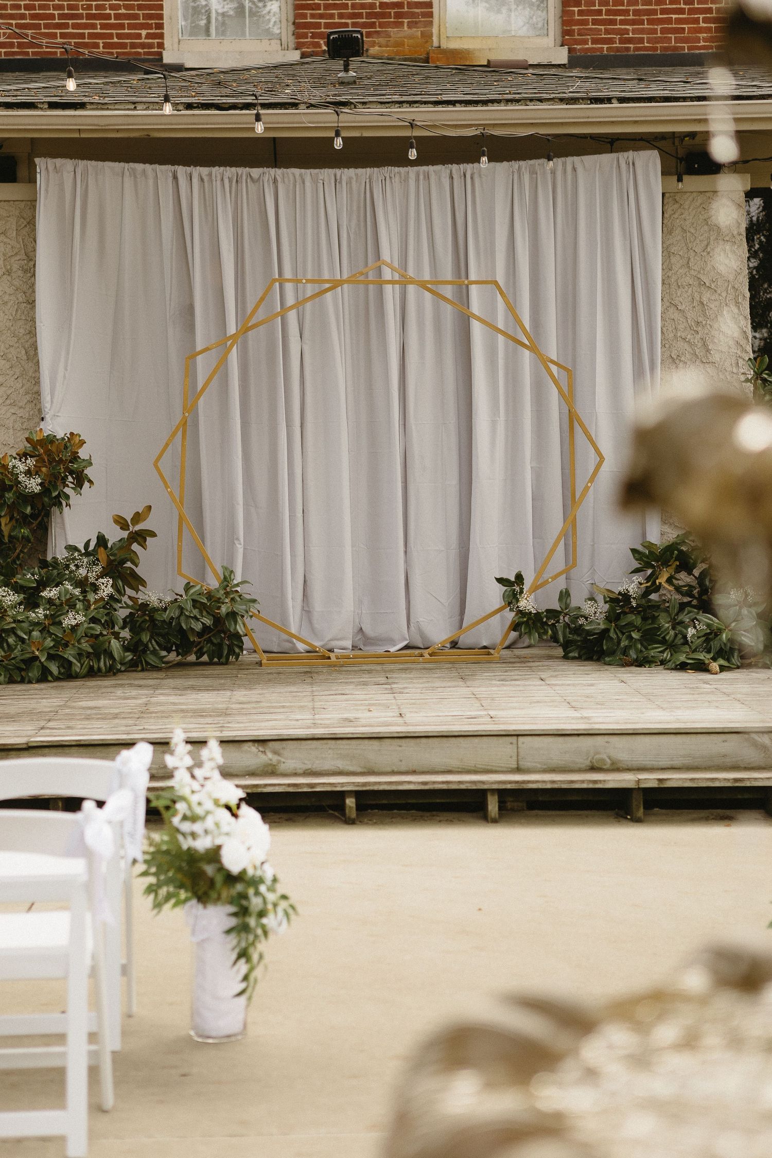 Wedding ceremony setup with white drapes and geometric gold arch against brick building.