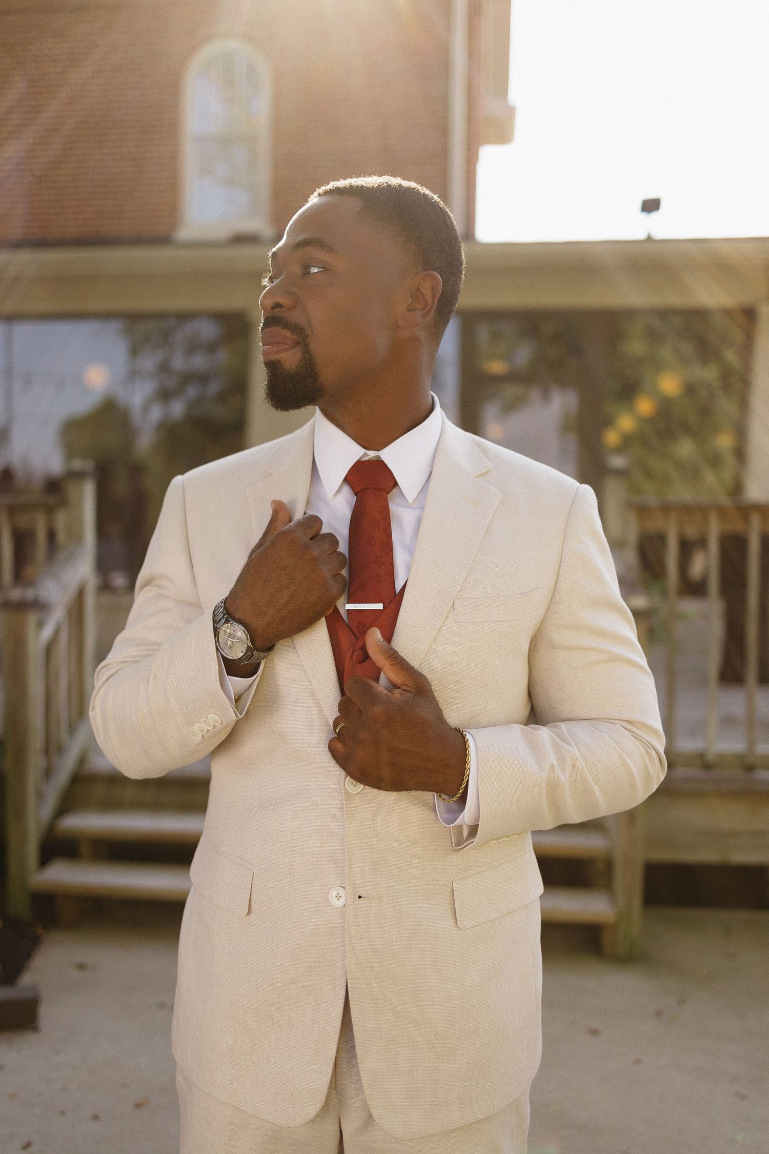 Groom in white suit and red tie adjusts his collar in warm sunlight.
