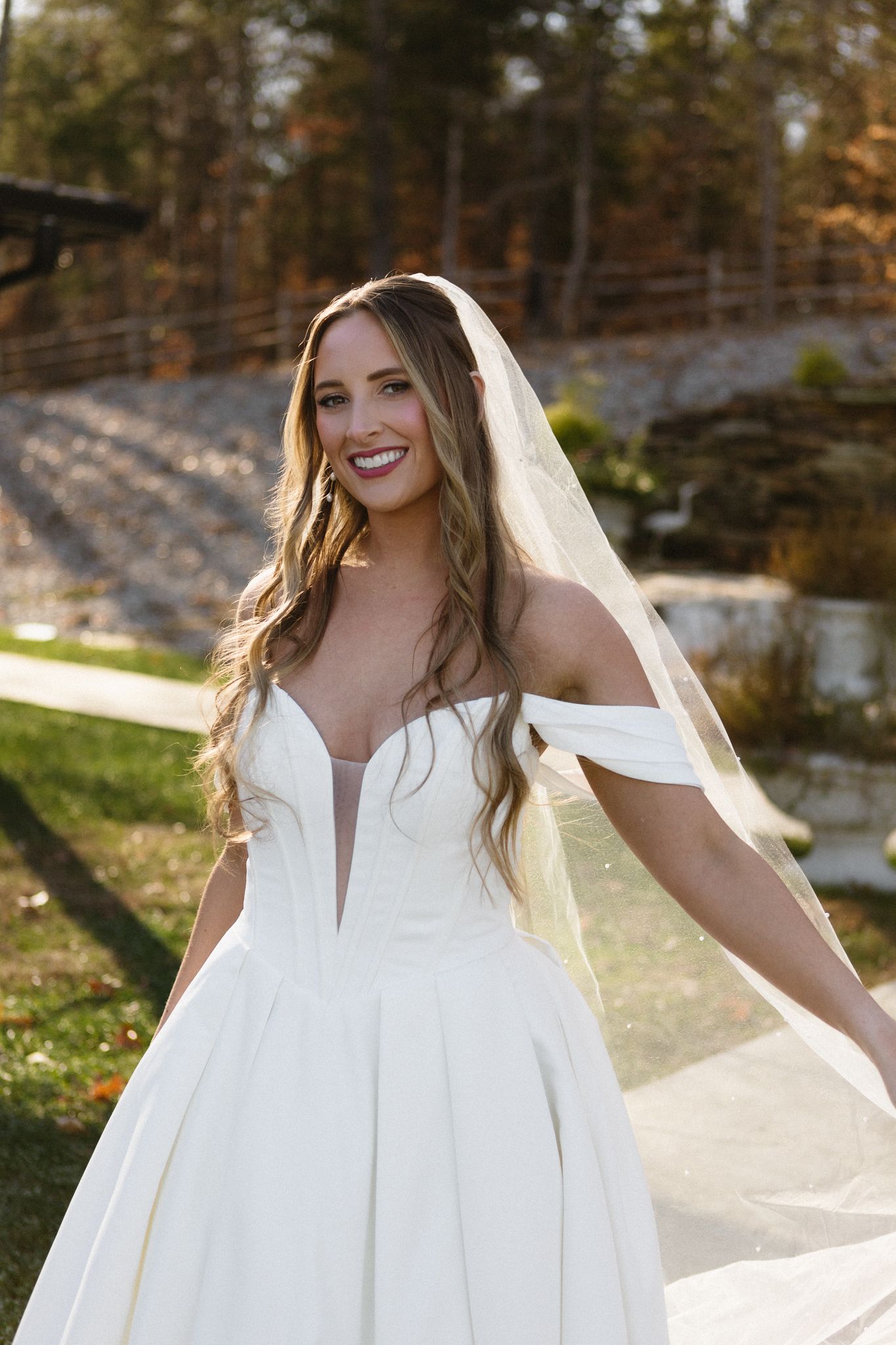Bride in off-shoulder wedding gown with flowing veil against autumn foliage.