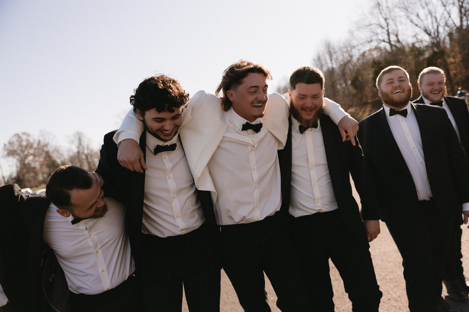 Groomsmen in black suits and white shirts stand together outdoors for a fun group wedding photo.
