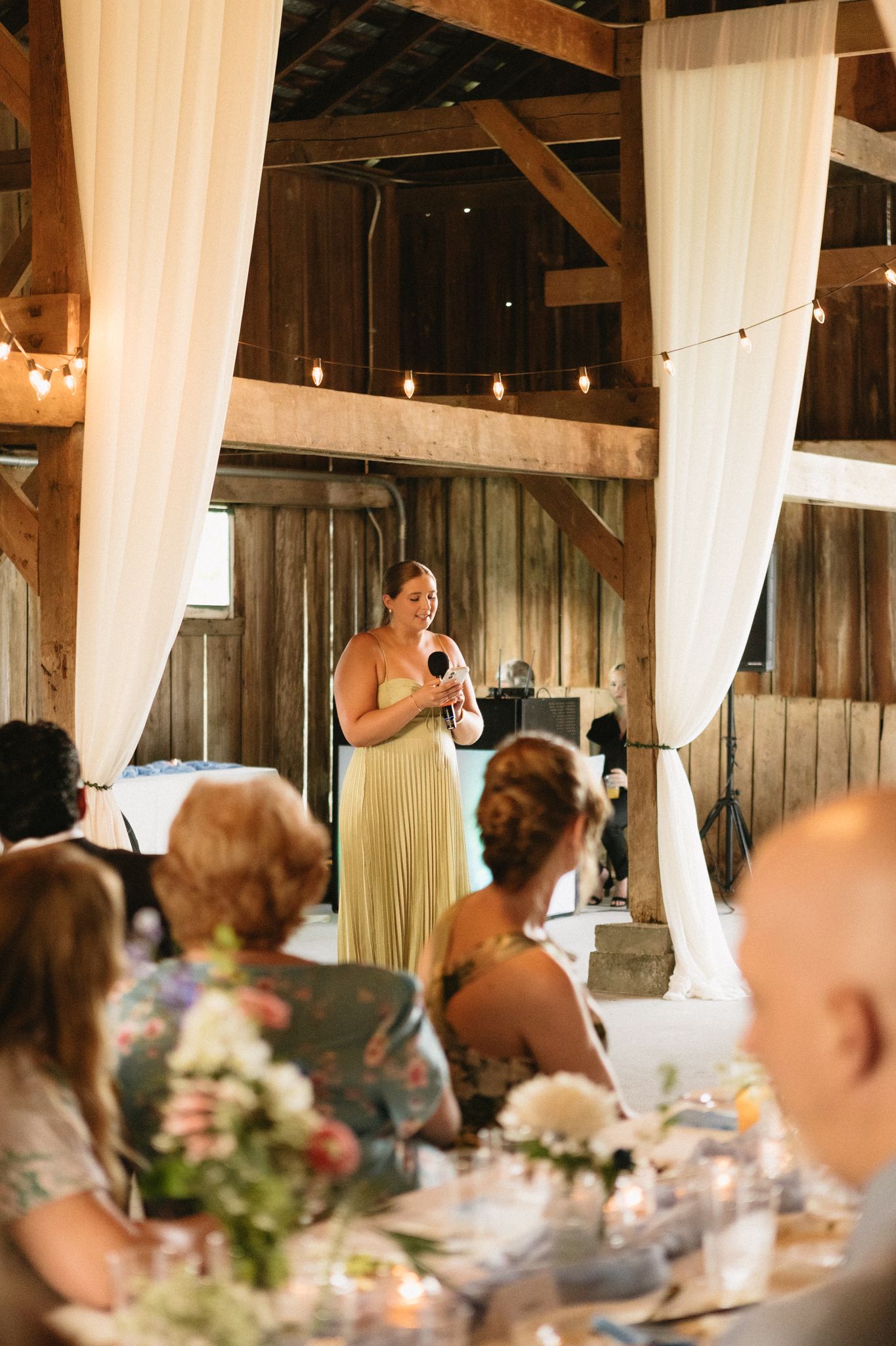 Wedding reception speeches in rustic barn venue decorated with white drapes and string lights.