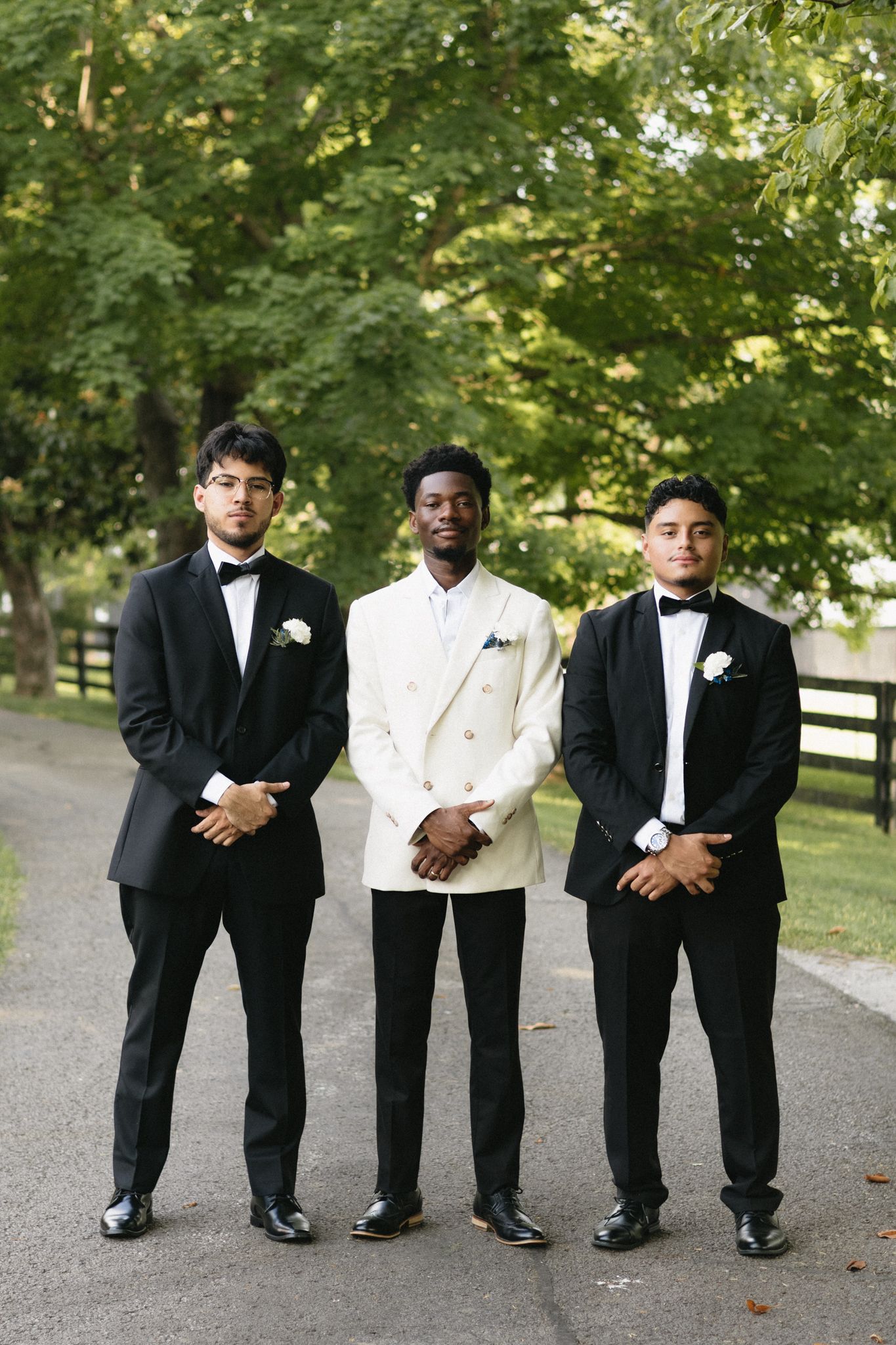 Groomsmen in classic tuxedos and white jacket pose together on tree-lined path.