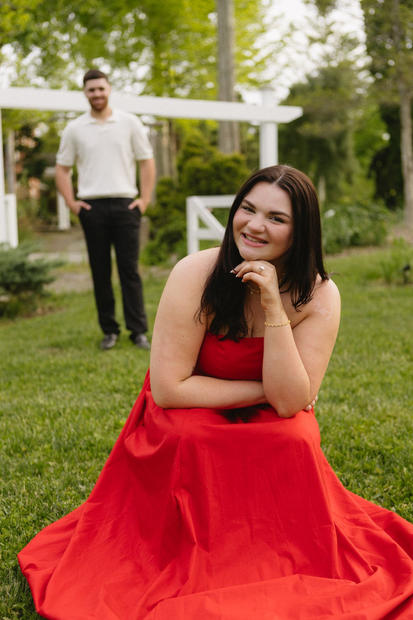 A playful outdoor portrait session with a woman in a red dress and a man standing in the background.