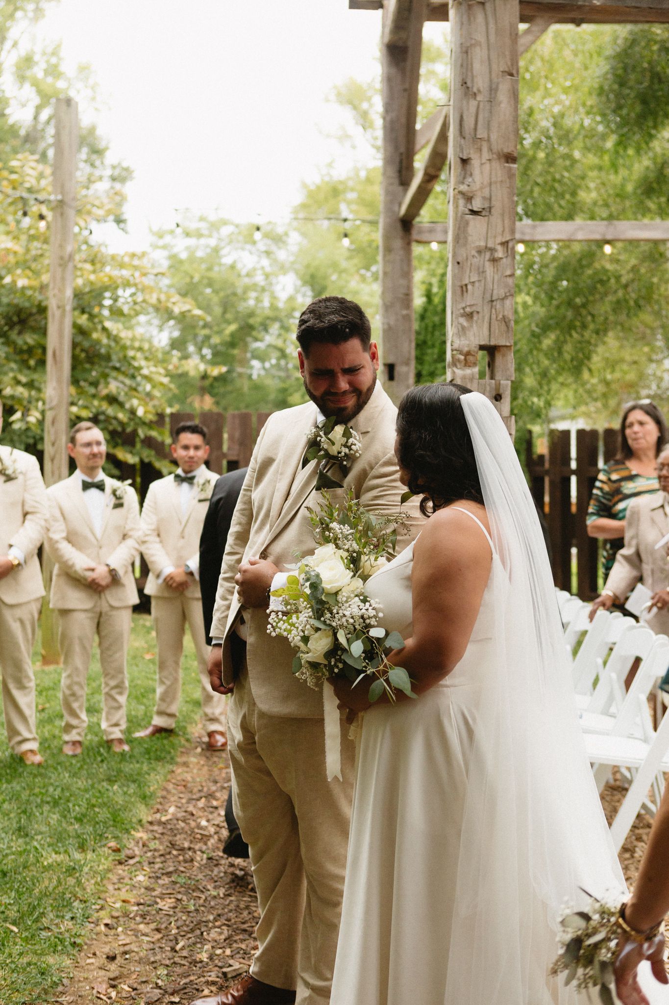 A wedding ceremony takes place under a rustic wooden arbor with white chairs and floral decorations in an outdoor setting.