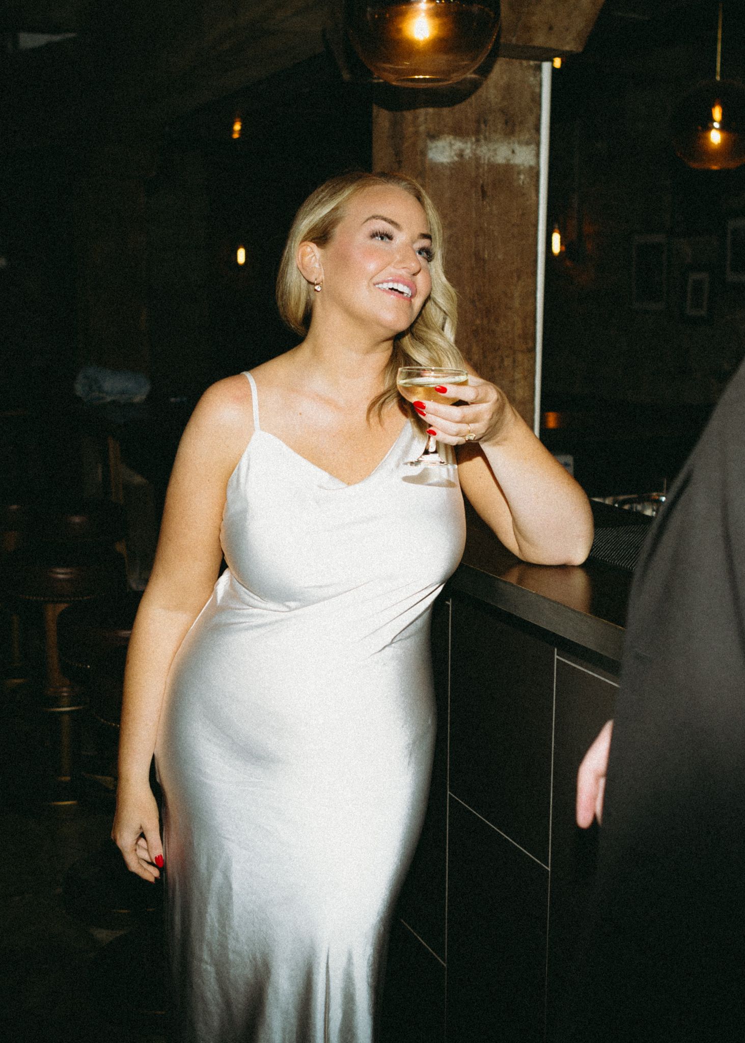 Someone in an elegant white cocktail dress raises a wine glass while enjoying drinks at a dimly lit bar setting.