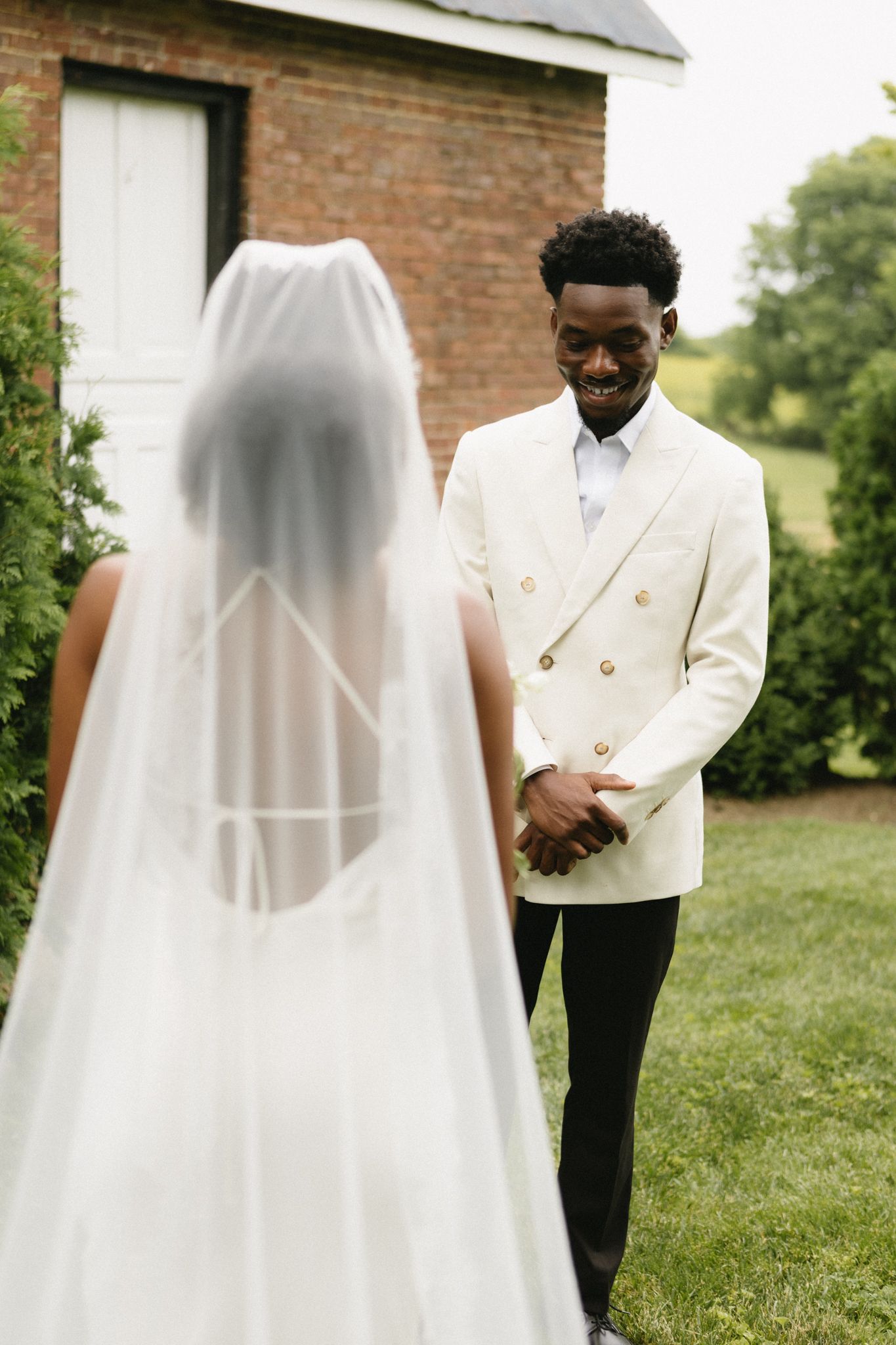 Wedding couple stands against brick building with flowing white veil and formal attire in natural light.