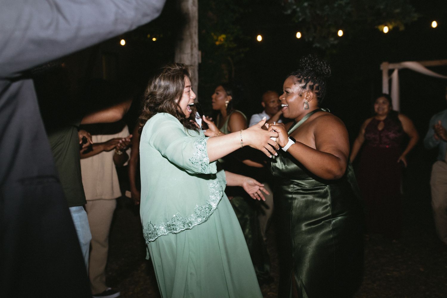 Two wedding guests in mint green and green satin dresses dance enthusiastically at an evening reception with string lights above.