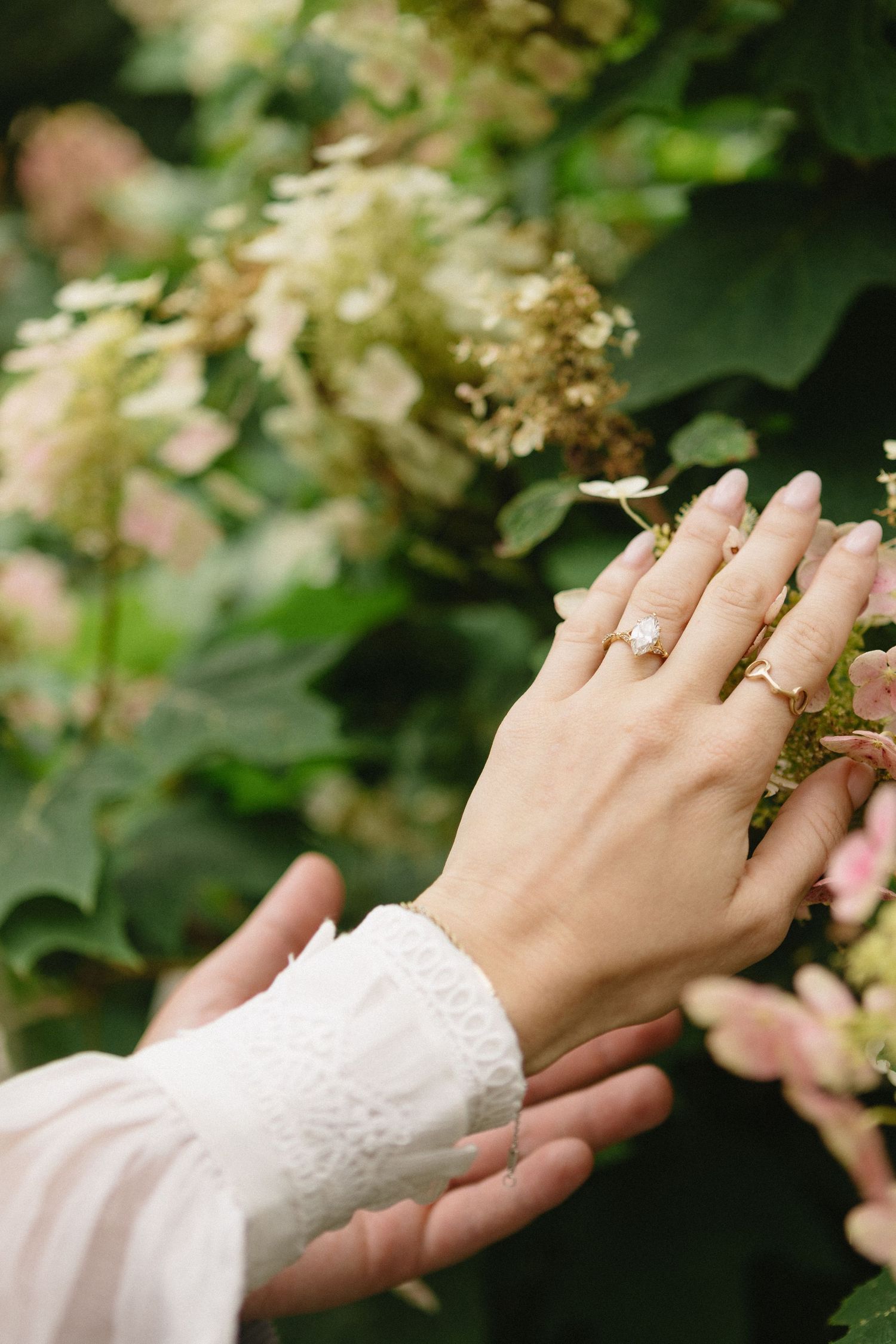 Close-up of womans hand displaying diamond engagement ring while touching blooming hydrangea flowers.