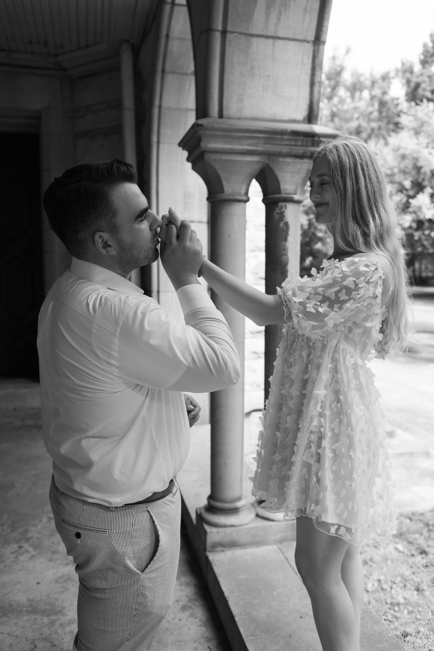 Black and white photo of a couple sharing a romantic moment near stone columns outdoors.
