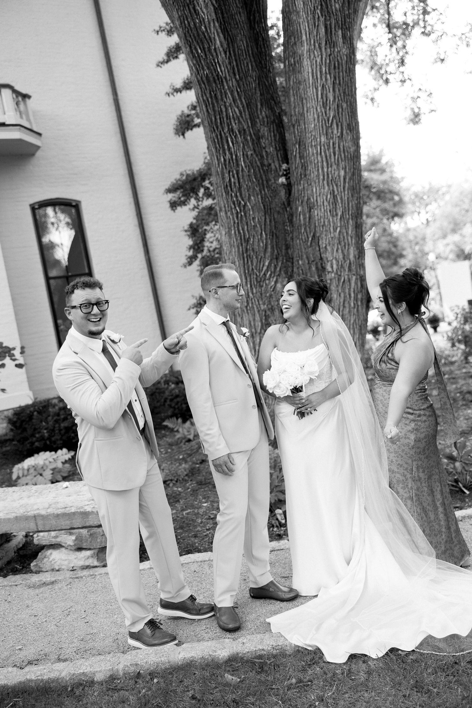 Black and white photo of bride and groom with wedding guests laughing outside a white building.