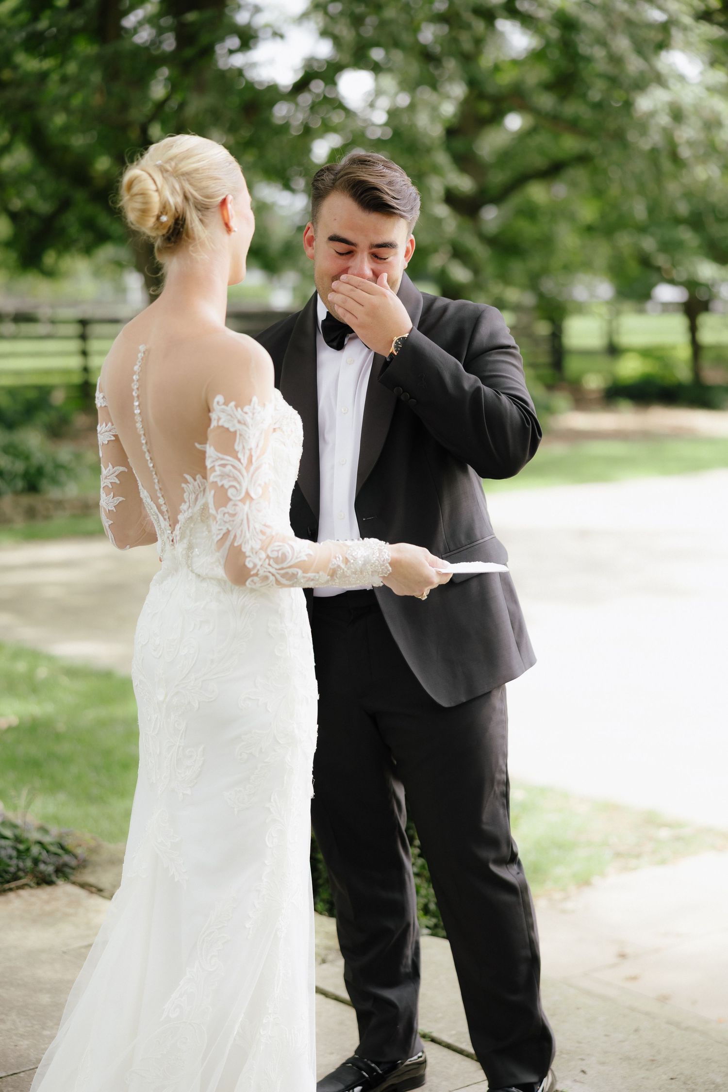 Emotional groom covering his mouth as bride in lace wedding dress shares vows outdoors.
