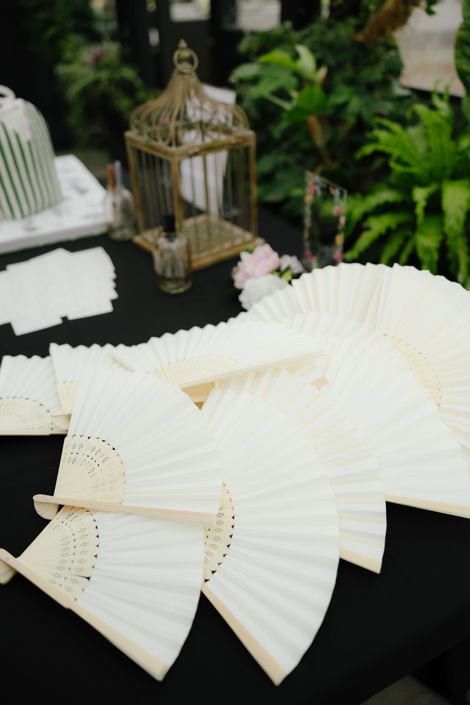 White decorative hand fans arranged on a black table beside a vintage gold birdcage at a wedding.