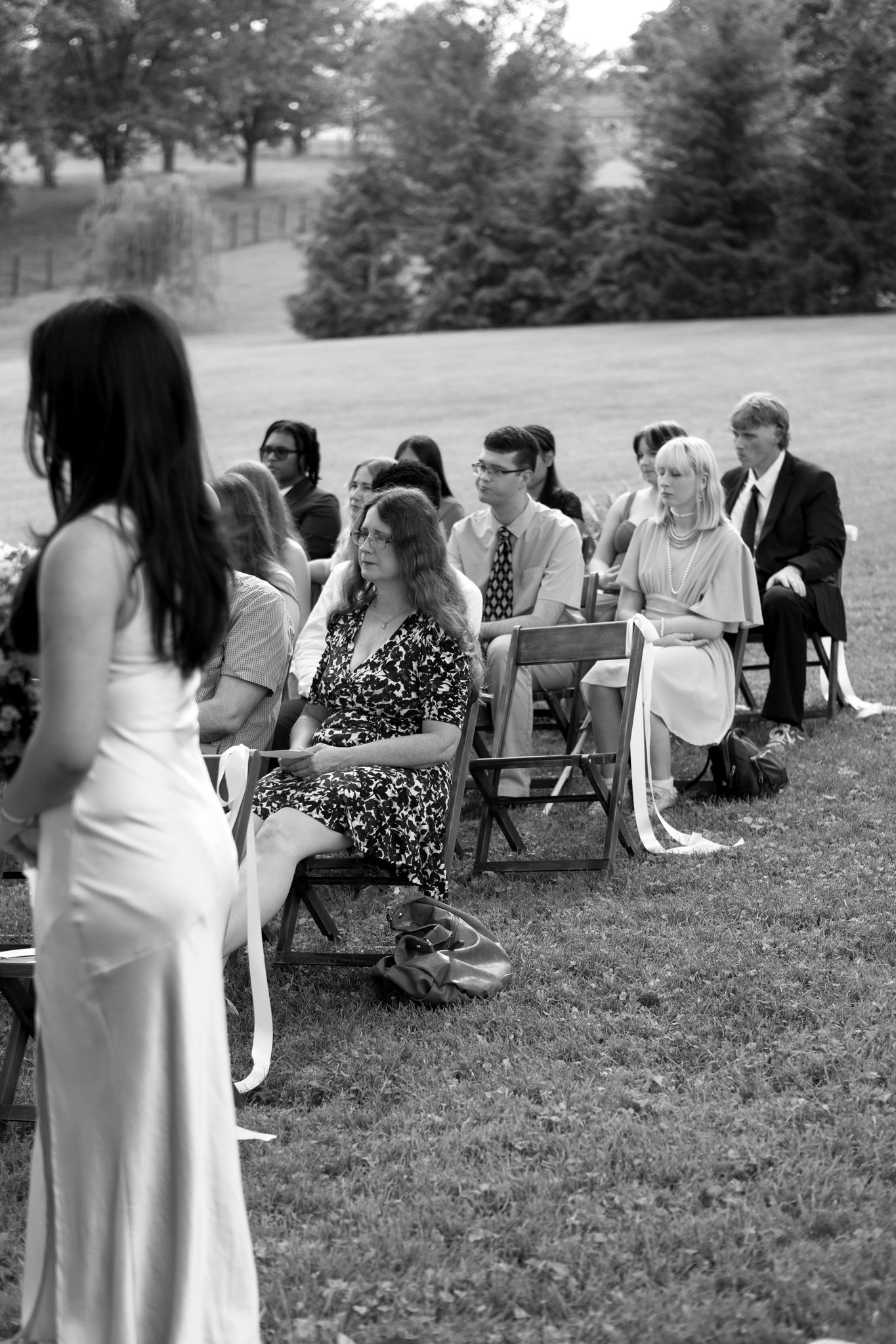Wedding guests seated on white chairs during an outdoor ceremony with trees in the background.