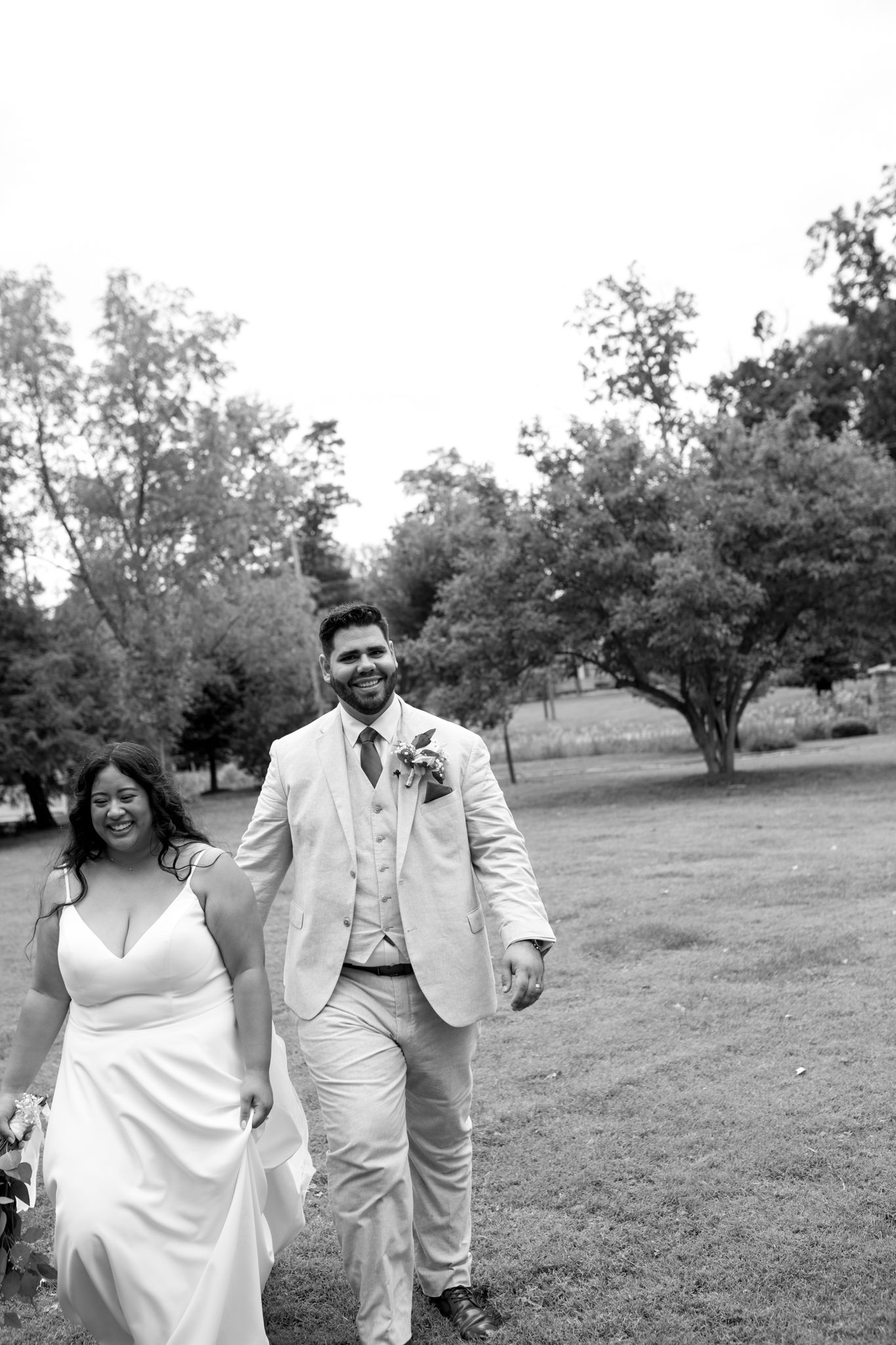 A black and white wedding photograph shows a couple walking together on a grassy lawn surrounded by trees.