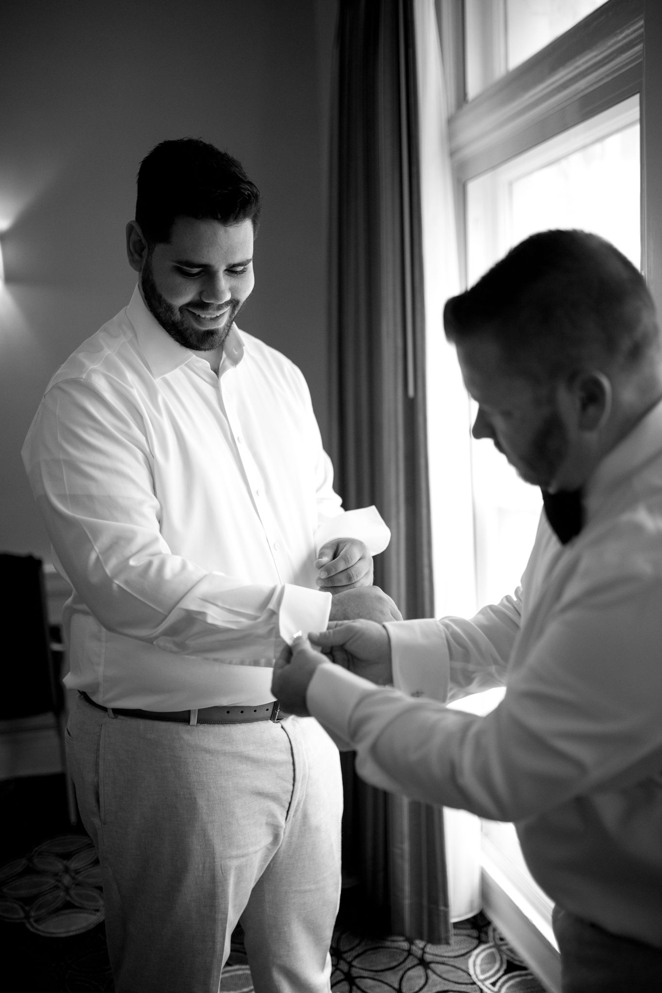 In a softly lit room by a window, a person in white helps adjust the cuffs on another's dress shirt during preparation.