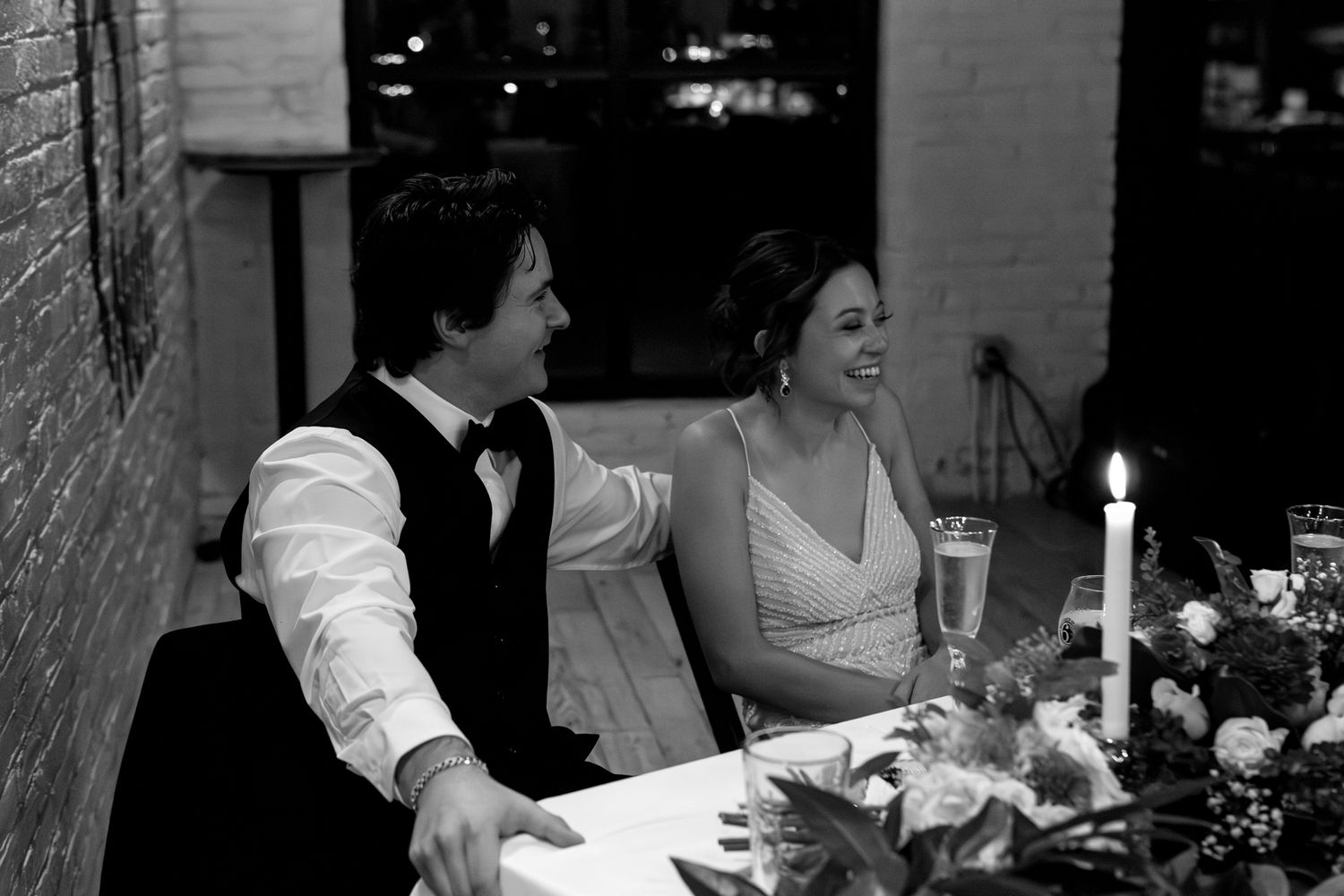 A couple sits together at an elegantly set candlelit dinner table during their wedding reception in black and white.